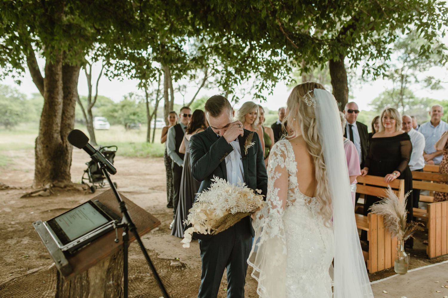 Wedding ceremony in progress under a natural tree canopy with wooden bench seating.