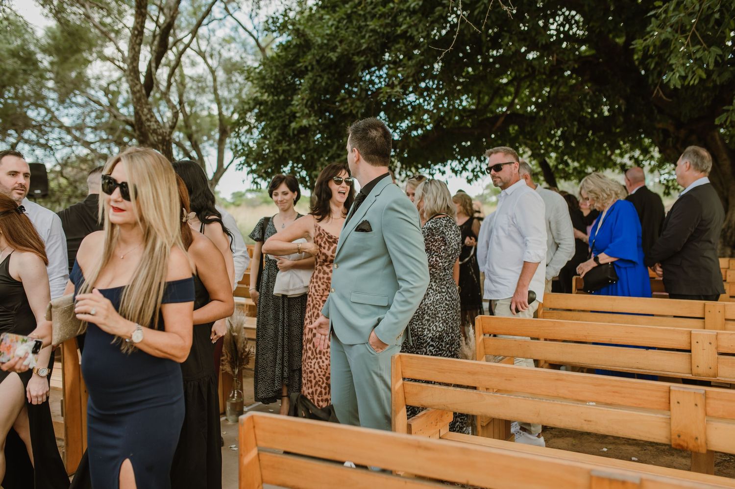 Wedding guests gather on wooden benches at an outdoor ceremony venue.