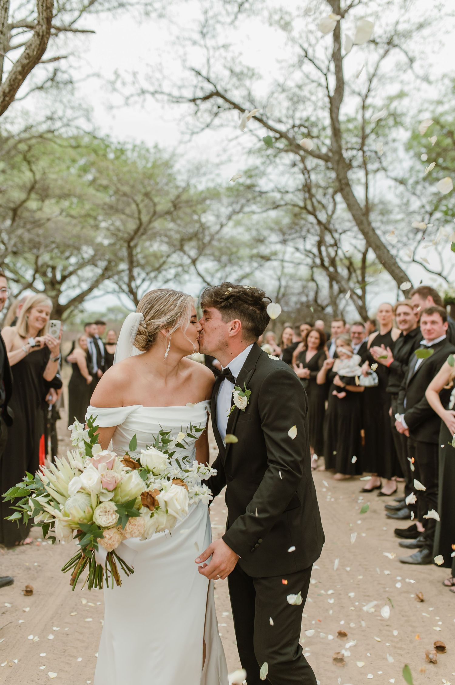 Romantic outdoor wedding ceremony with guests throwing confetti under tree canopy.
