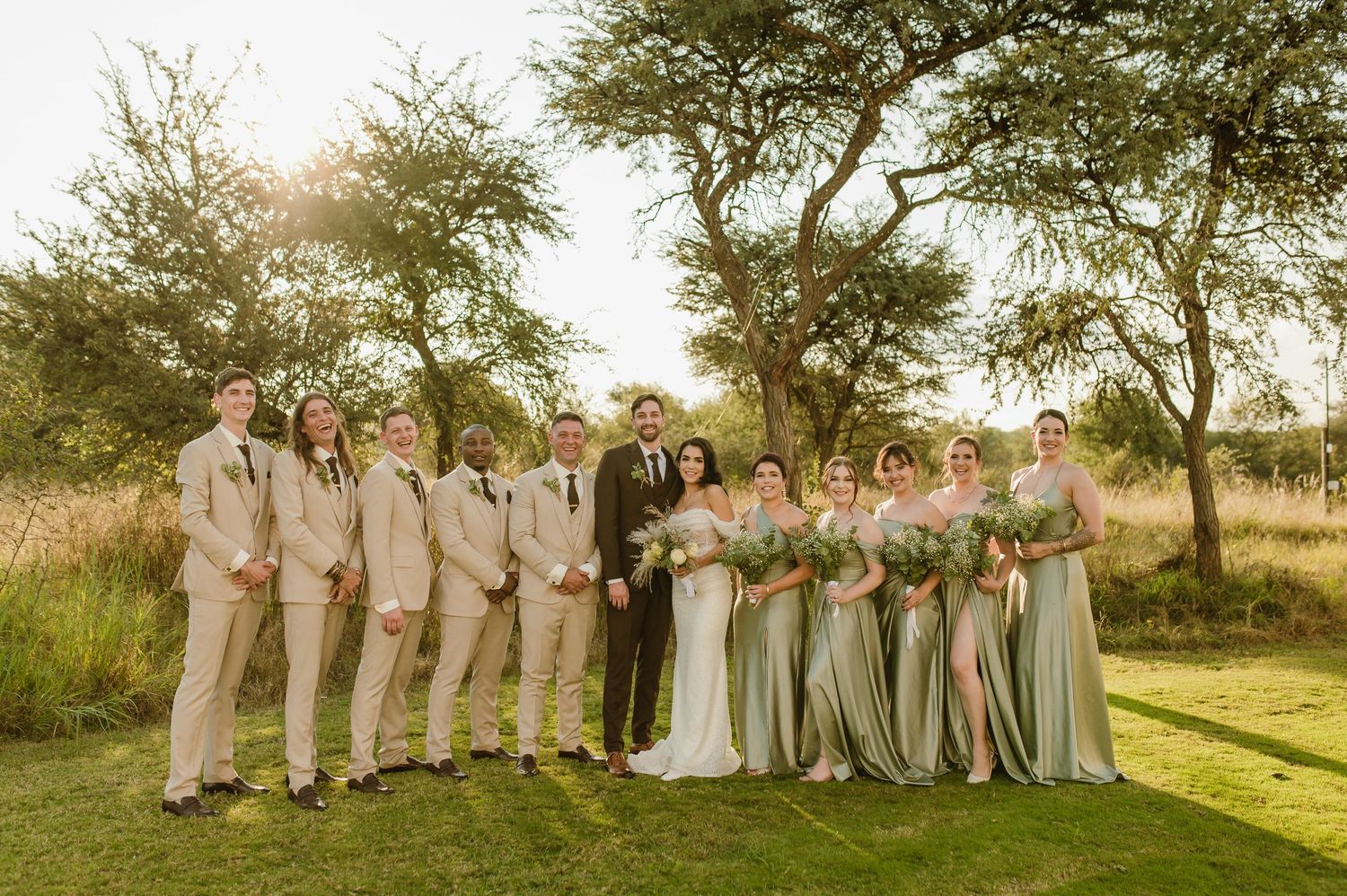 Wedding party poses together in sage green dresses and tan suits against natural landscape.