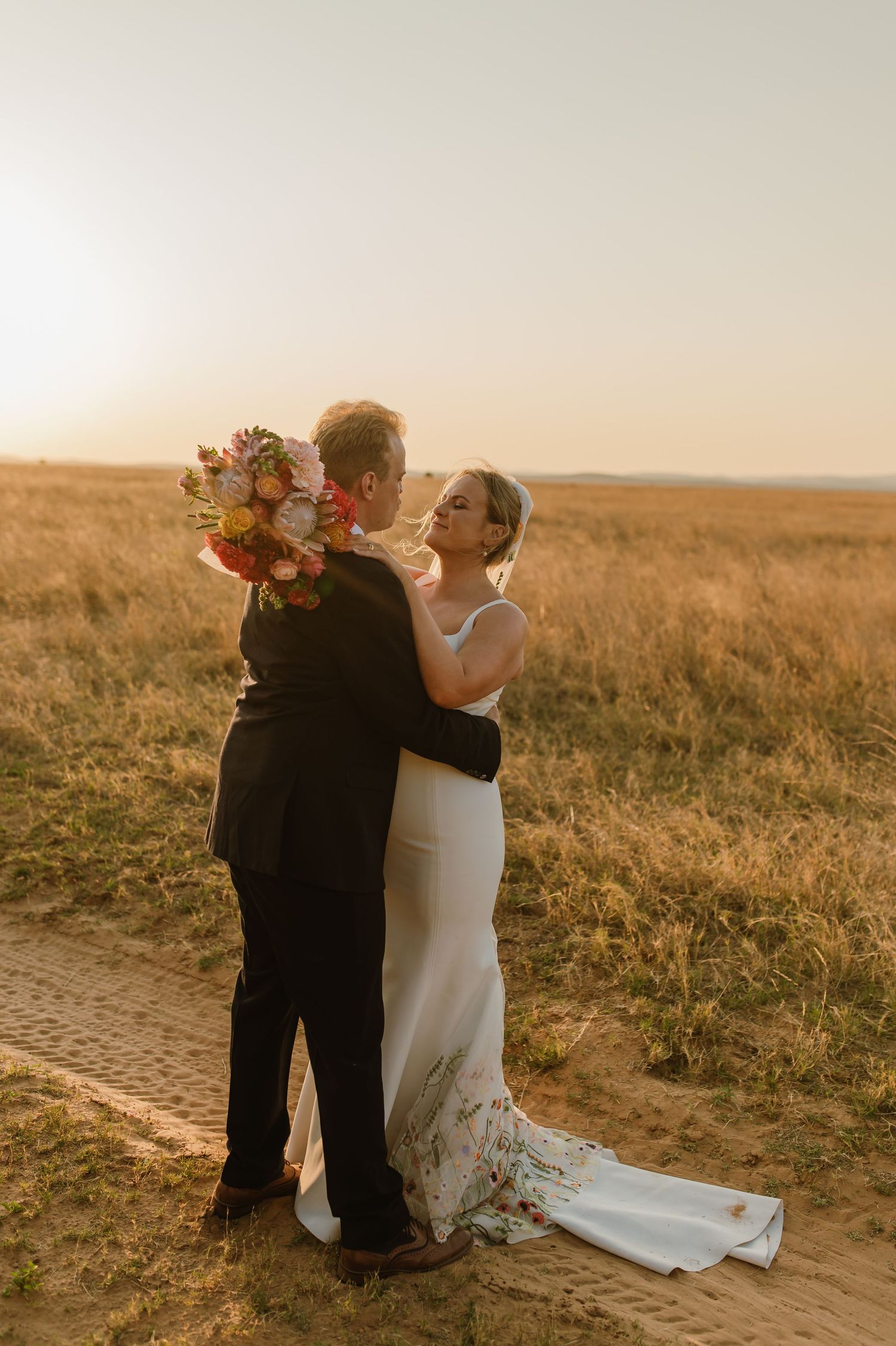 A couple embraces on a dirt path during their golden hour wedding photoshoot.