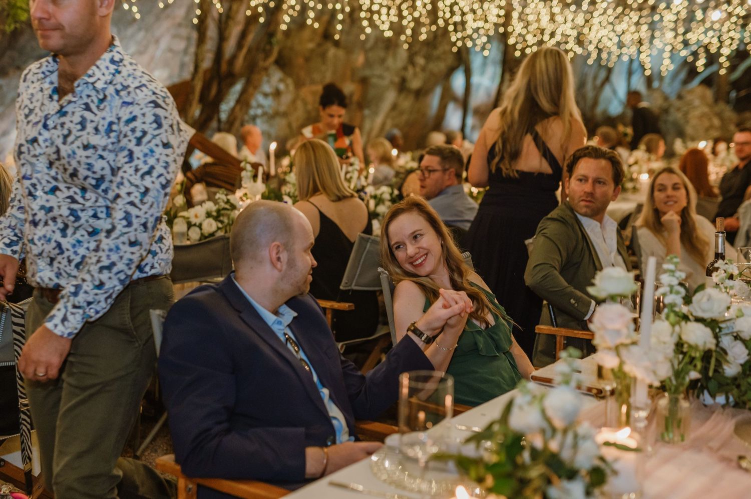 Wedding guests enjoying dinner at decorated tables under twinkling fairy lights in an outdoor setting.