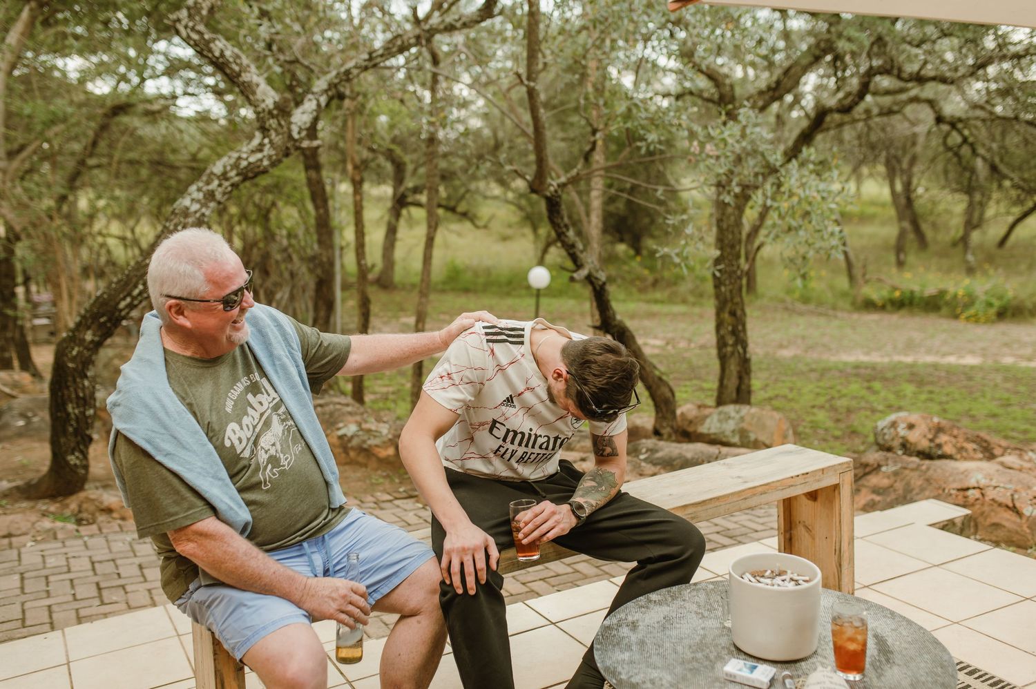 Friends socializing on a stone patio under a wooden pergola in a natural setting.