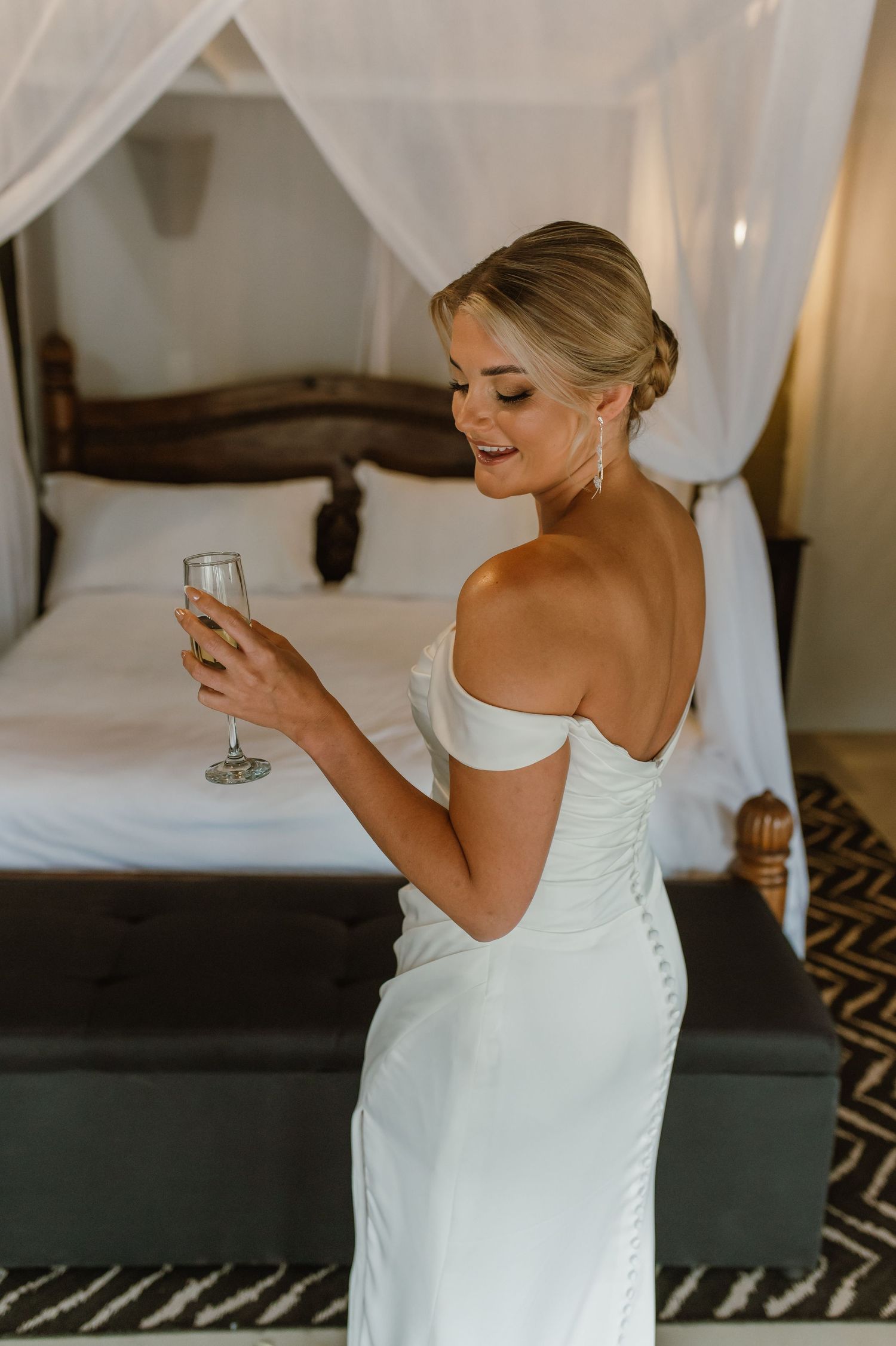 Bride in fitted white dress smiles while holding champagne glass near draped canopy bed.