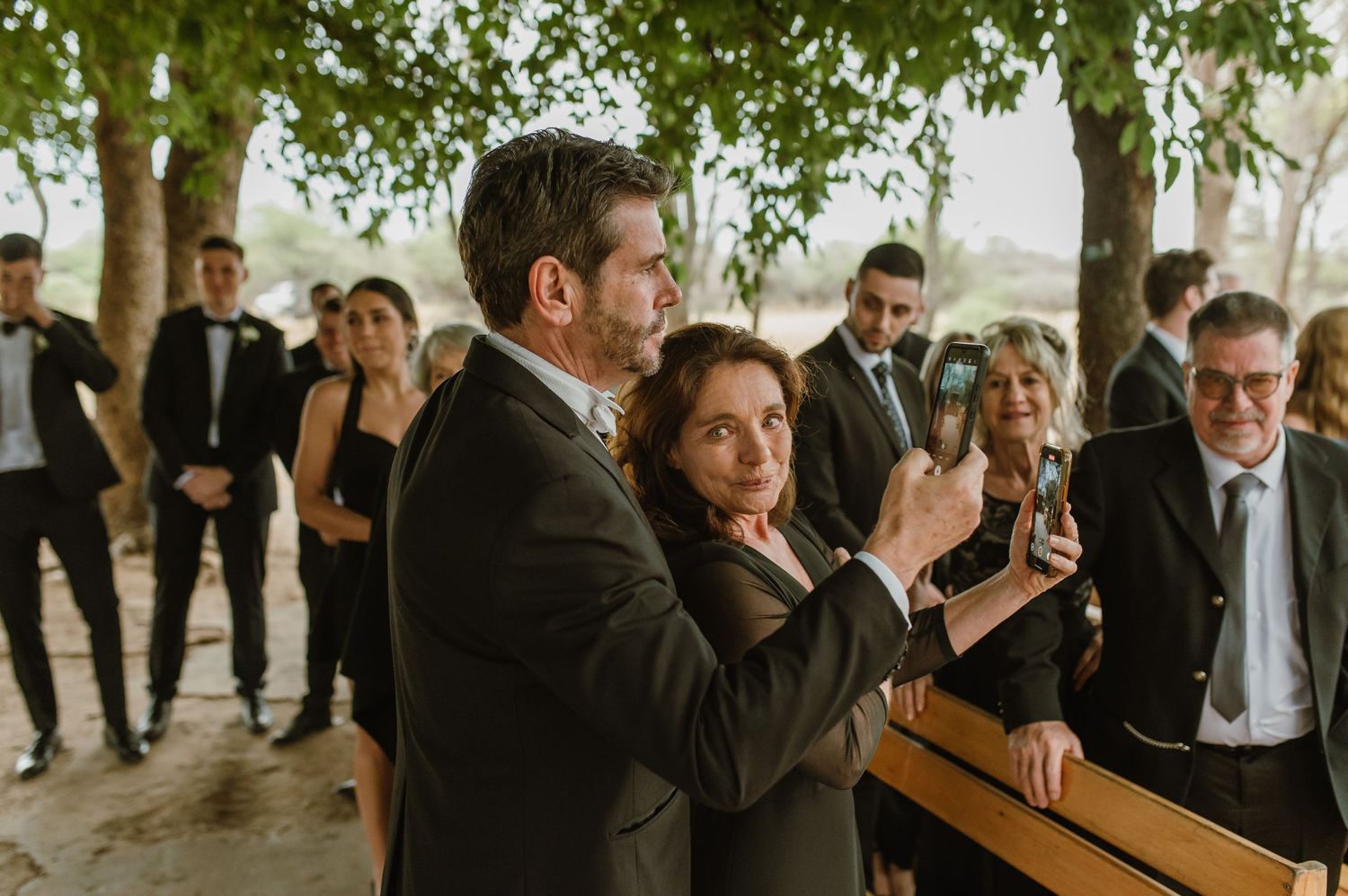 Wedding guests in formal attire gather under shaded trees during outdoor ceremony.