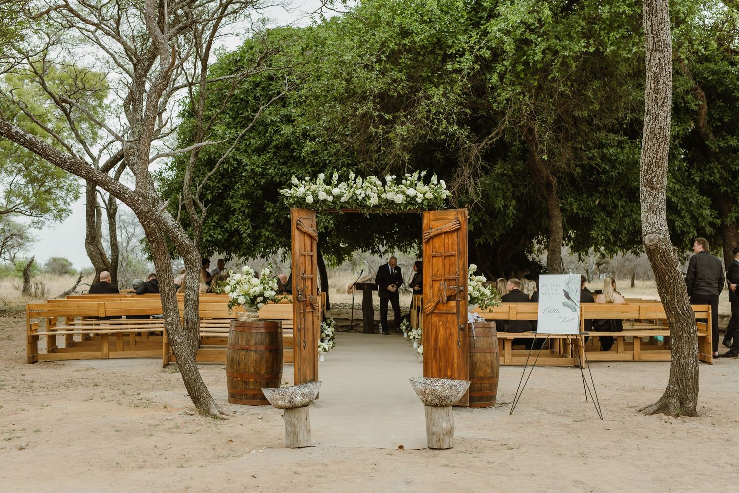 Wooden bench seating arranged outdoors under shady trees for a wedding ceremony.