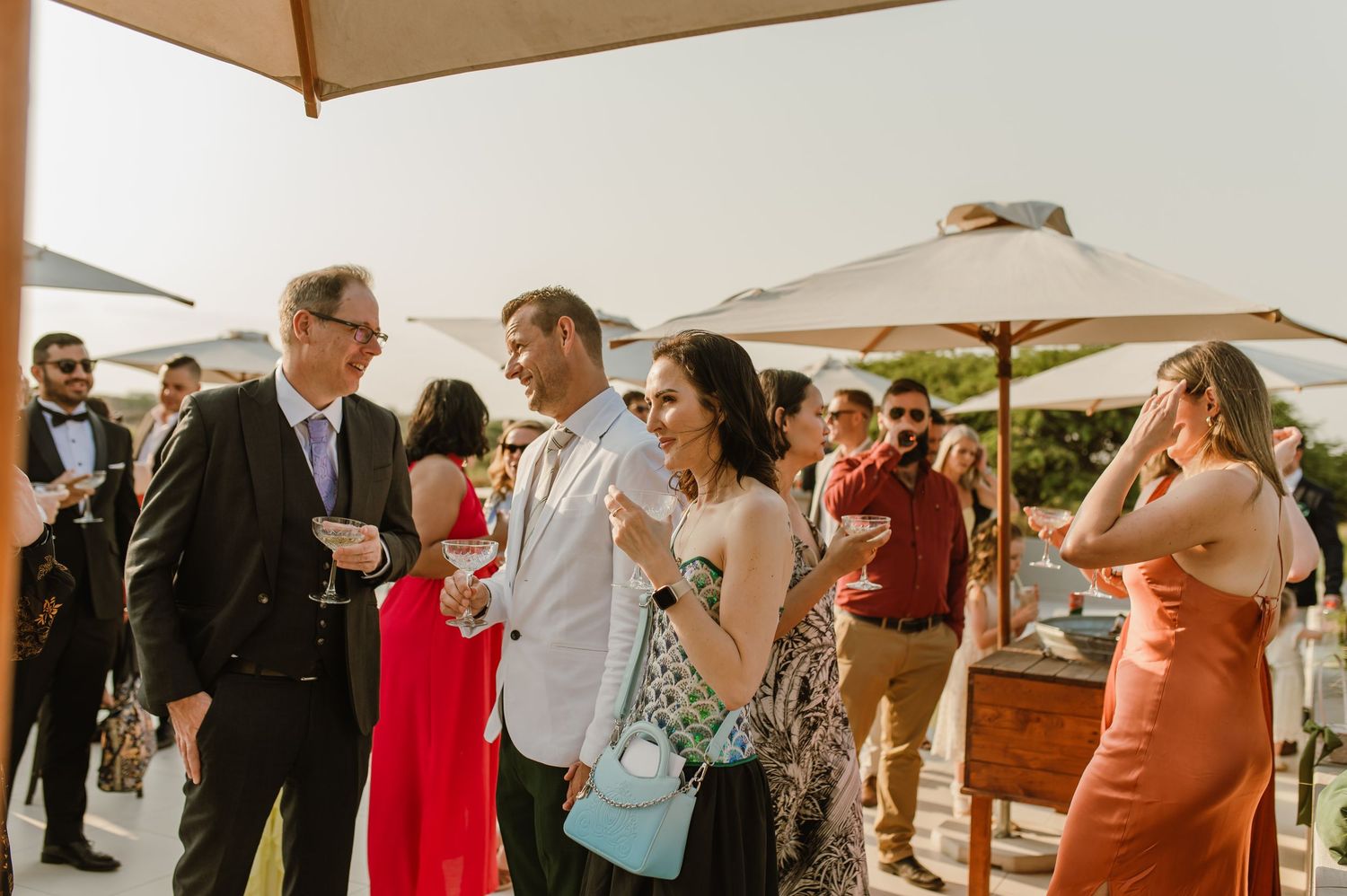 Large group of wedding guests socializing on an outdoor patio under tan umbrellas.