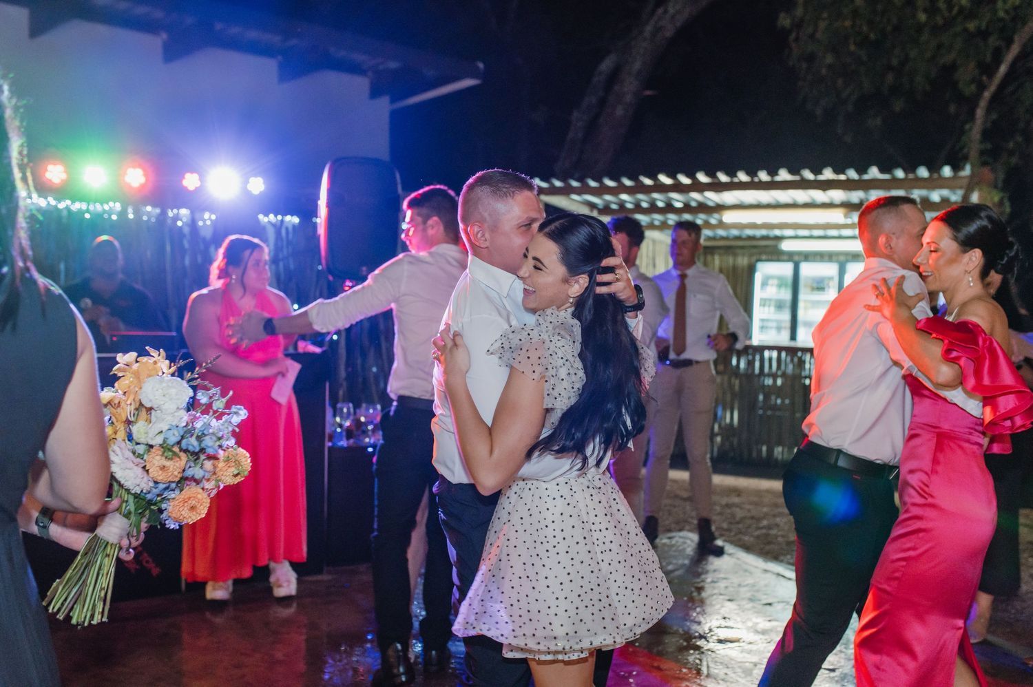 Bride in polka dot dress dances with groom while guests celebrate at outdoor wedding reception.