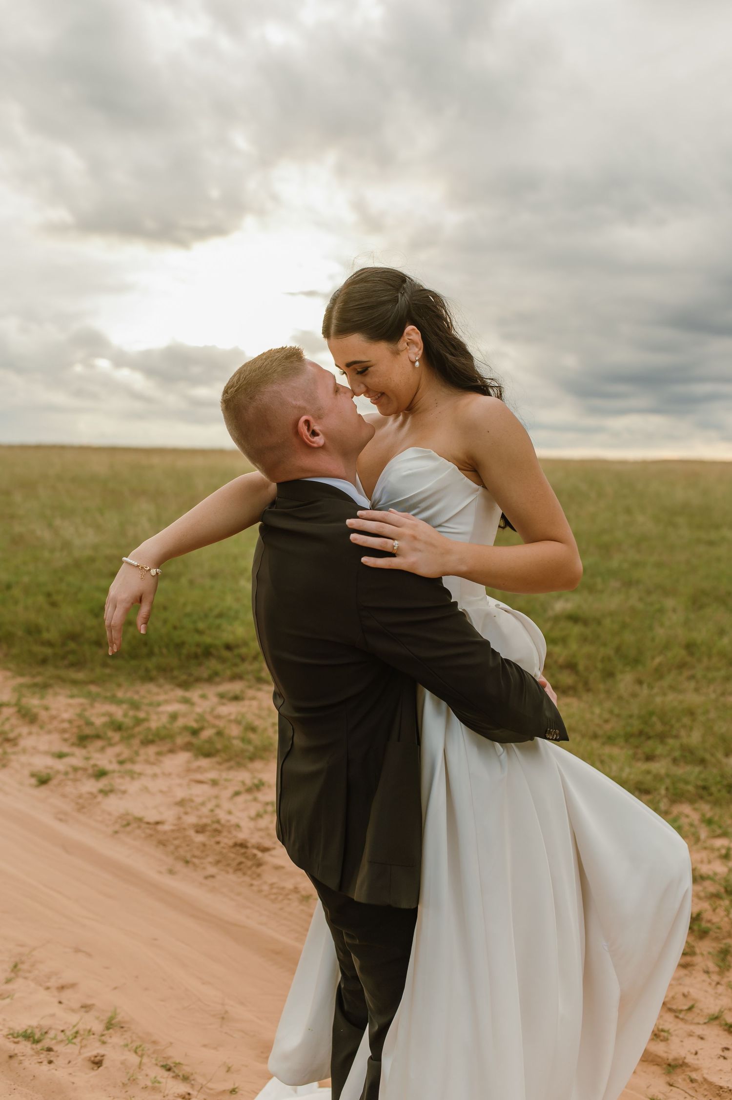 Groom lifting bride in white gown on sandy path with green fields and cloudy sky backdrop.