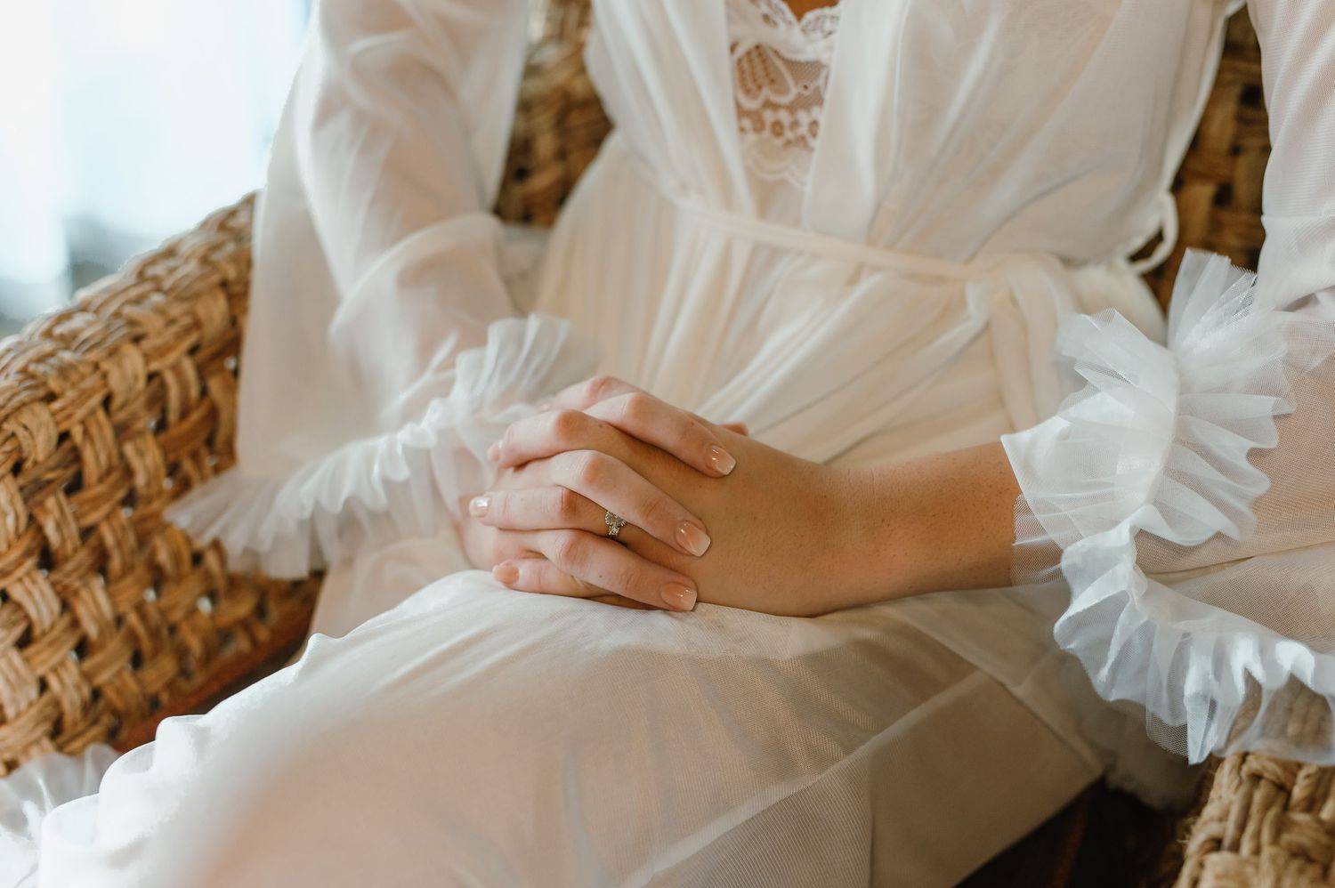Bride in white lace robe with hands folded in lap seated in wicker chair.