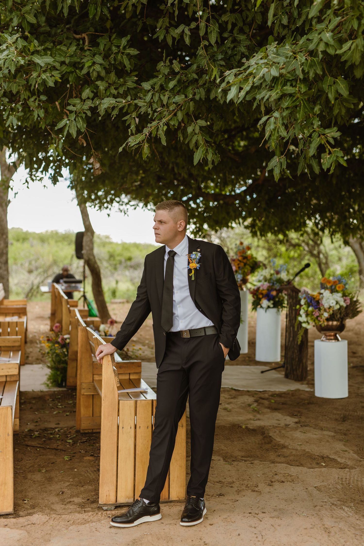 Groom in black suit with floral boutonniere stands beside wooden pew at outdoor wedding ceremony under green trees.