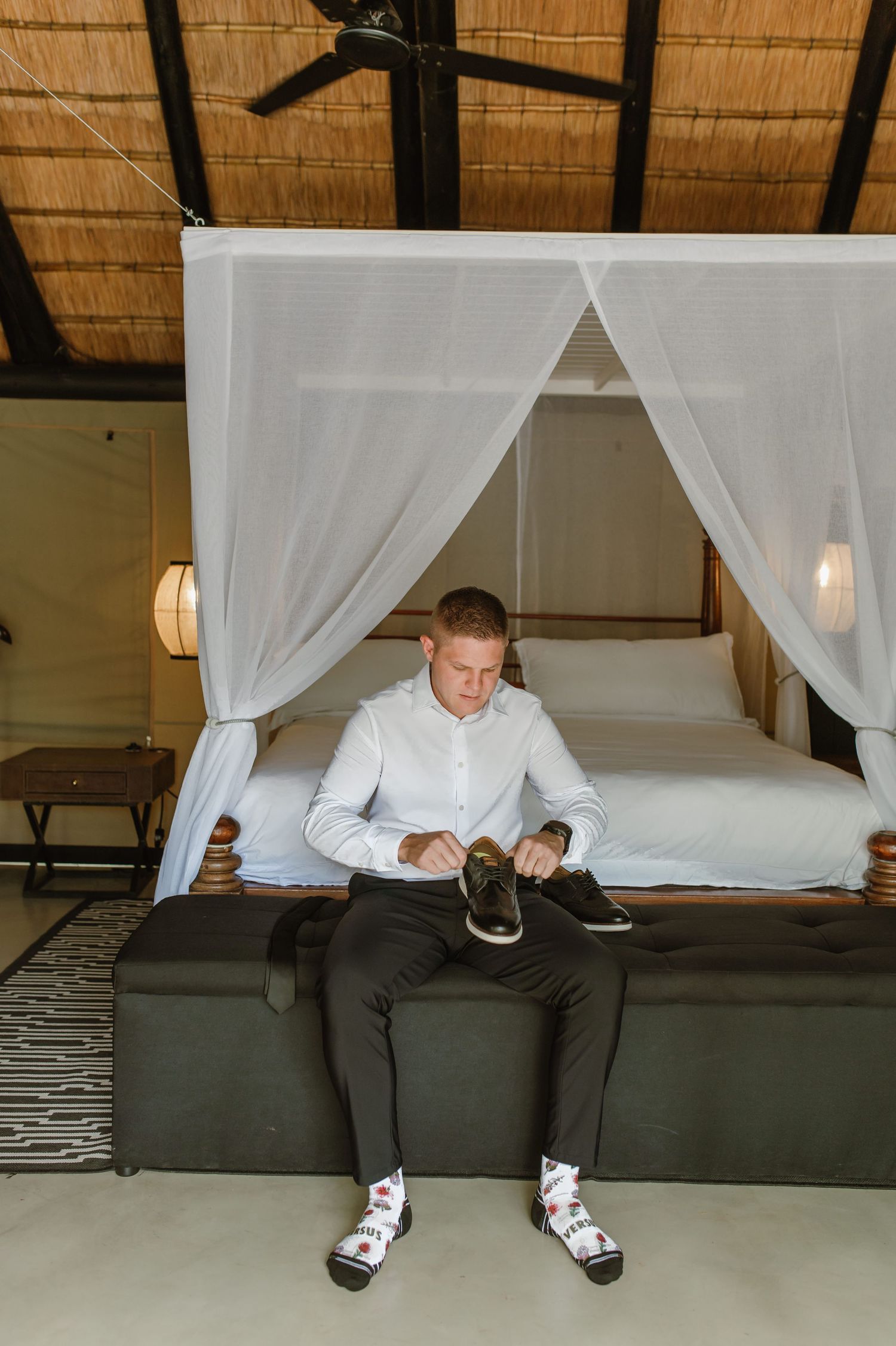 Groom sitting on bed putting on dress shoes with fun patterned socks in a safari lodge bedroom.