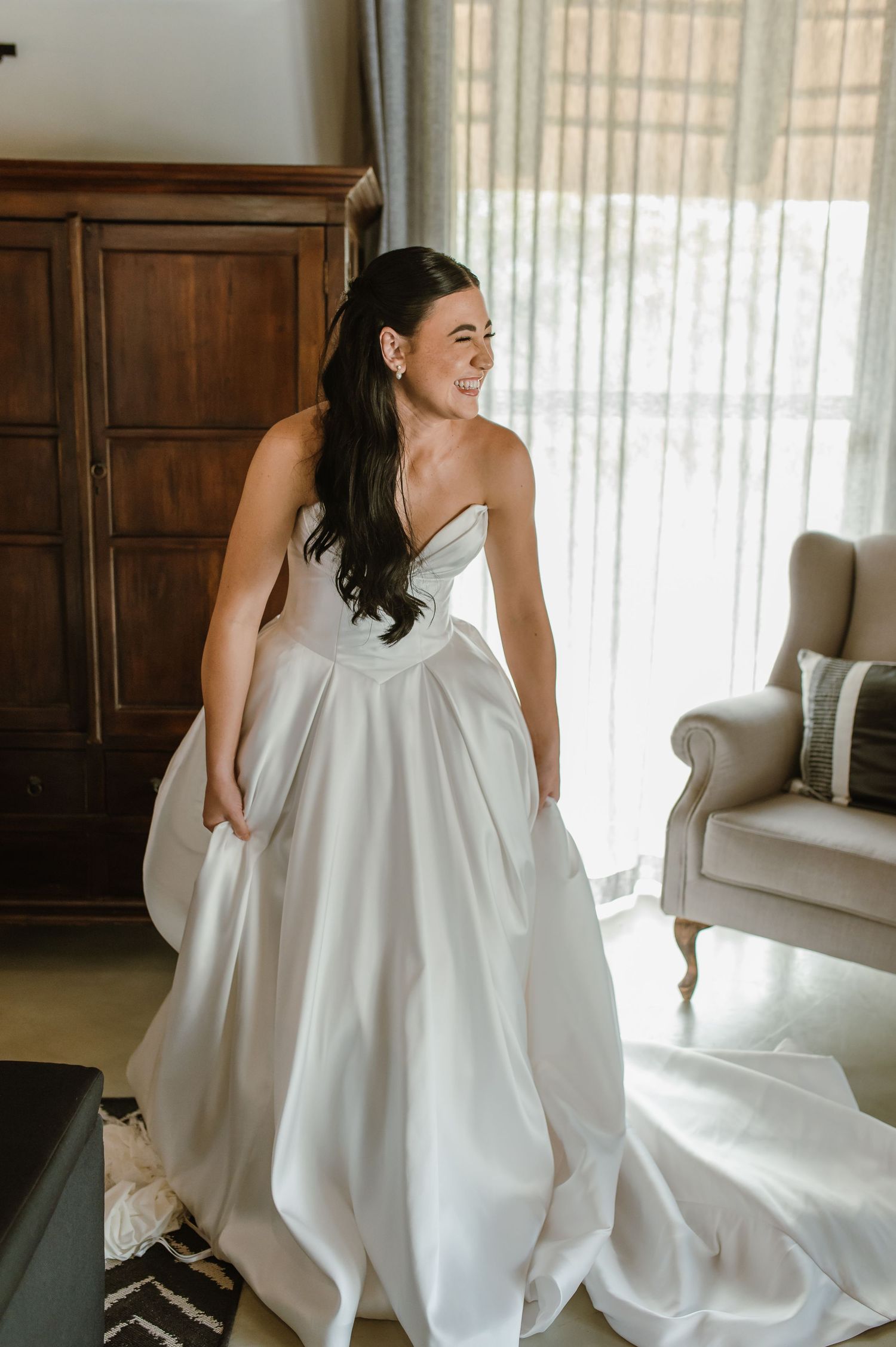 Smiling bride in strapless white satin ball gown holding her skirt in a sunlit room.