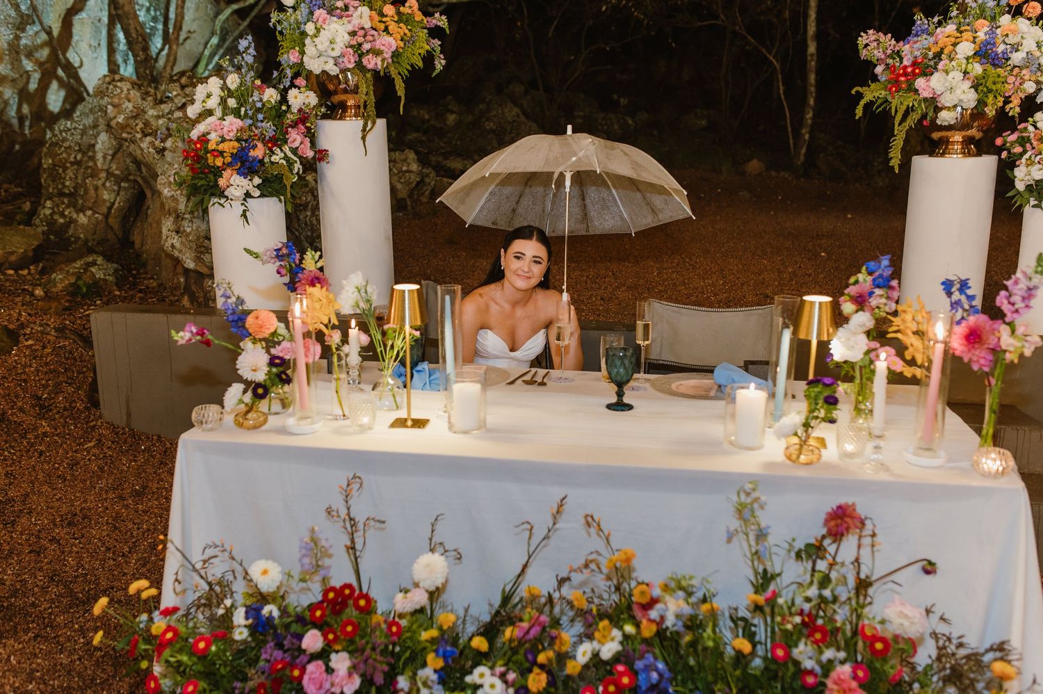 Bride sitting alone at floral-decorated sweetheart table holding umbrella surrounded by colorful wildflowers at dusk.