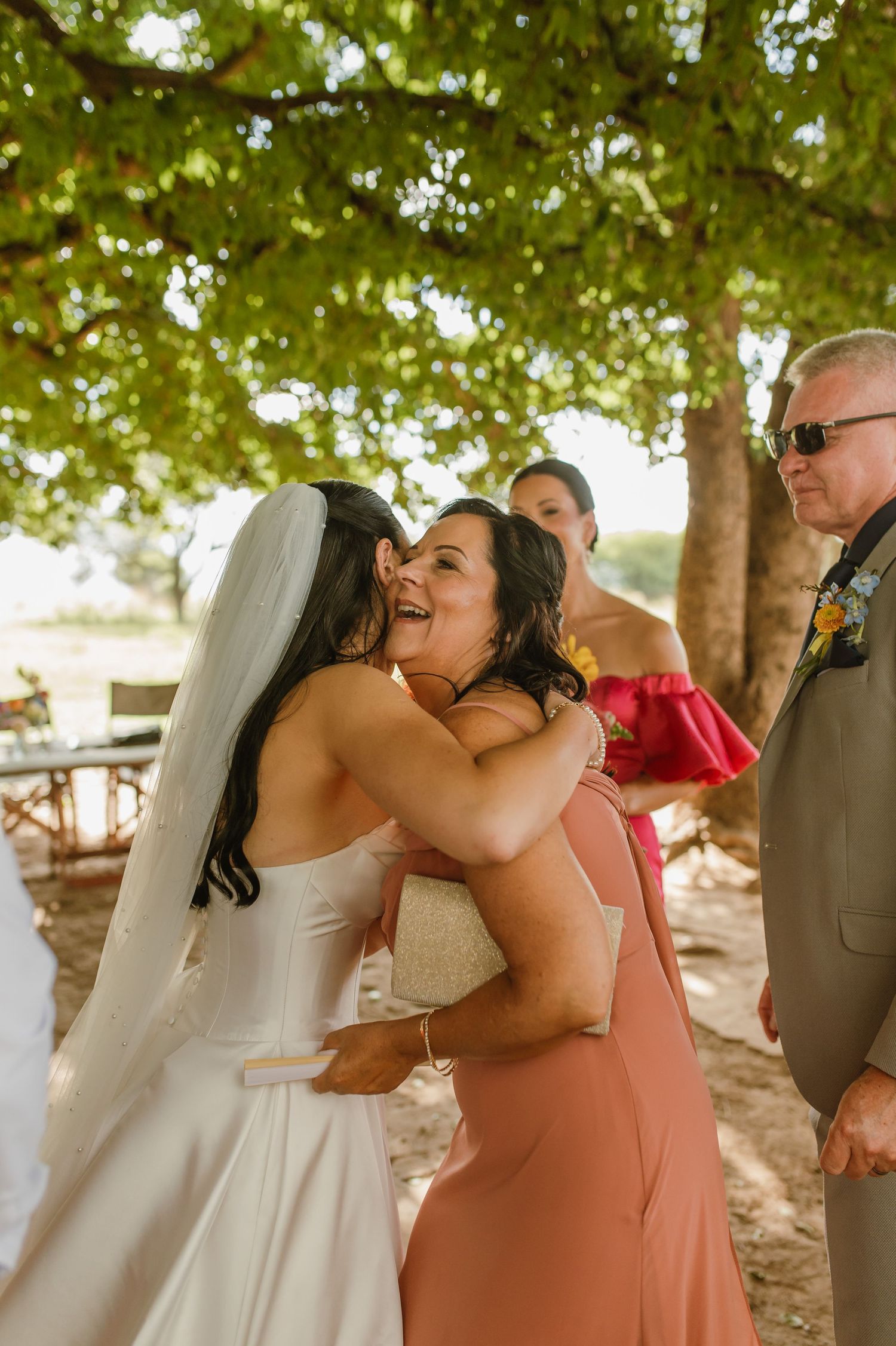 Bride in white wedding dress and veil sharing a warm embrace with a smiling female wedding guest.