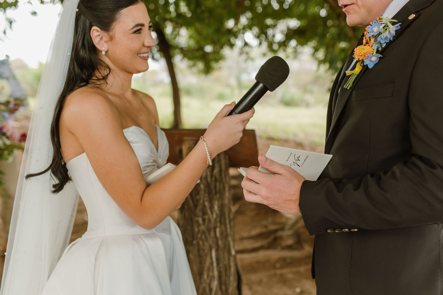 Smiling bride in strapless white gown holding microphone while groom reads wedding vows at outdoor ceremony.