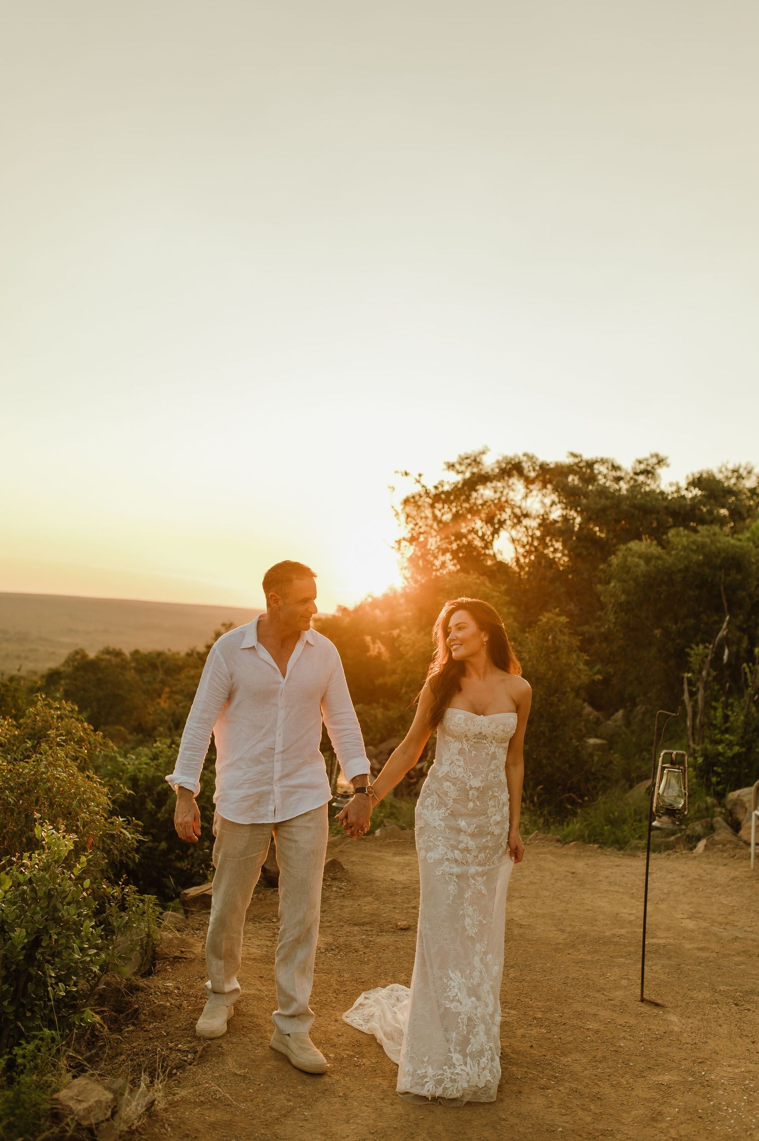 Newlyweds holding hands walking at sunset in outdoor wedding attire.