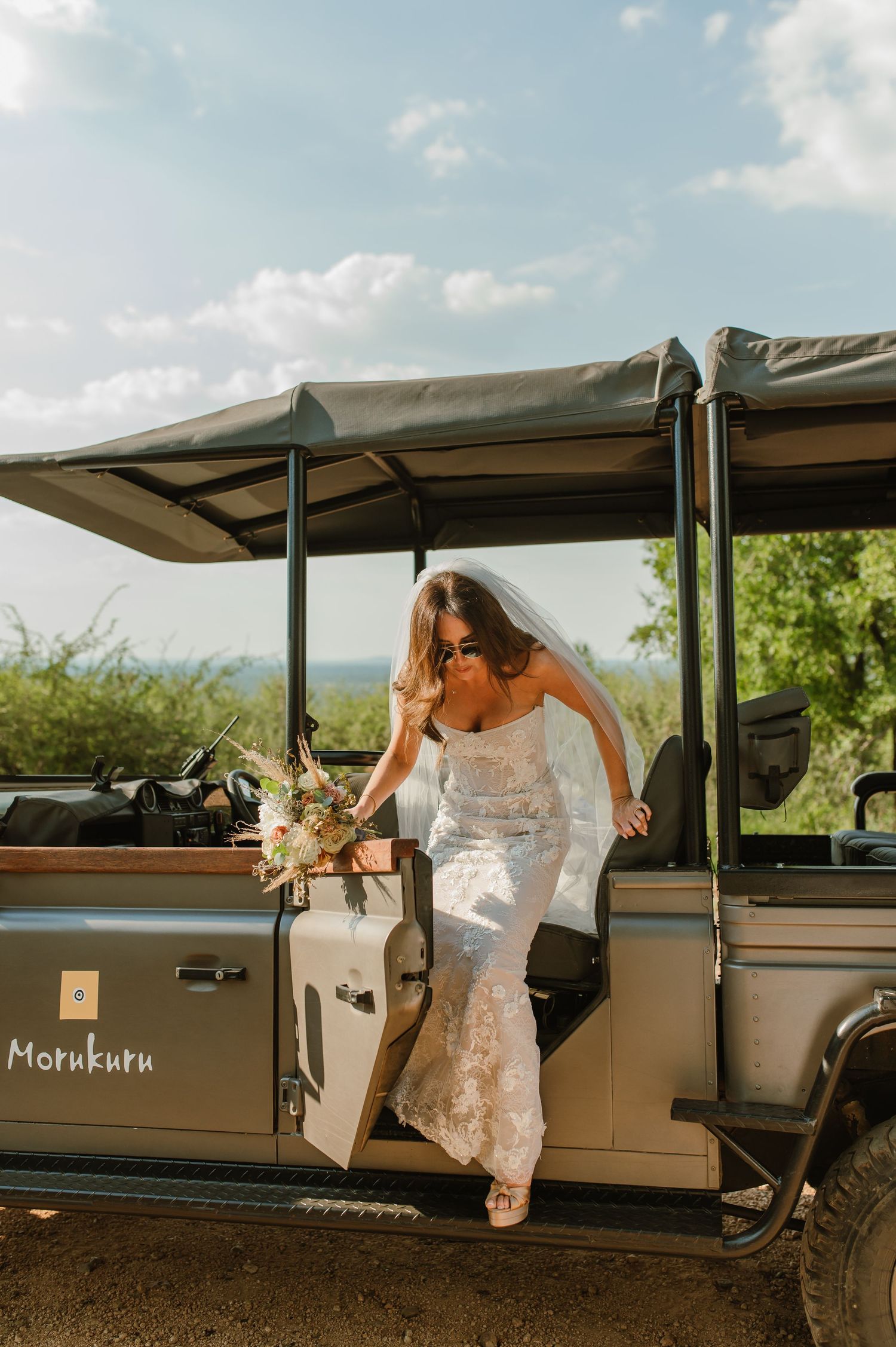 Bride in lace wedding dress and veil stepping off a Morukuru safari jeep holding a bouquet in the African bush.