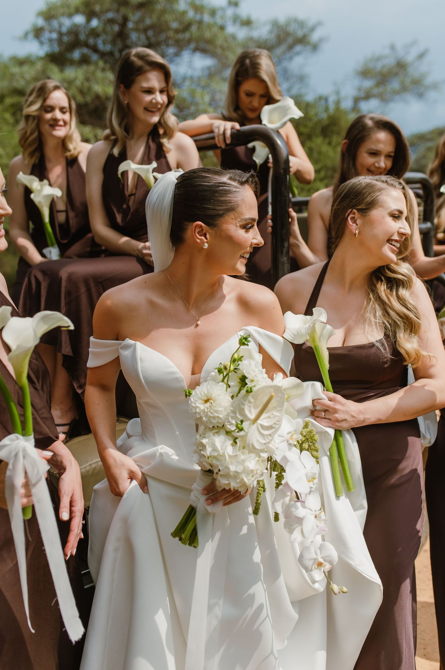 Wedding party in brown and white dresses holding orchid bouquets outdoors.