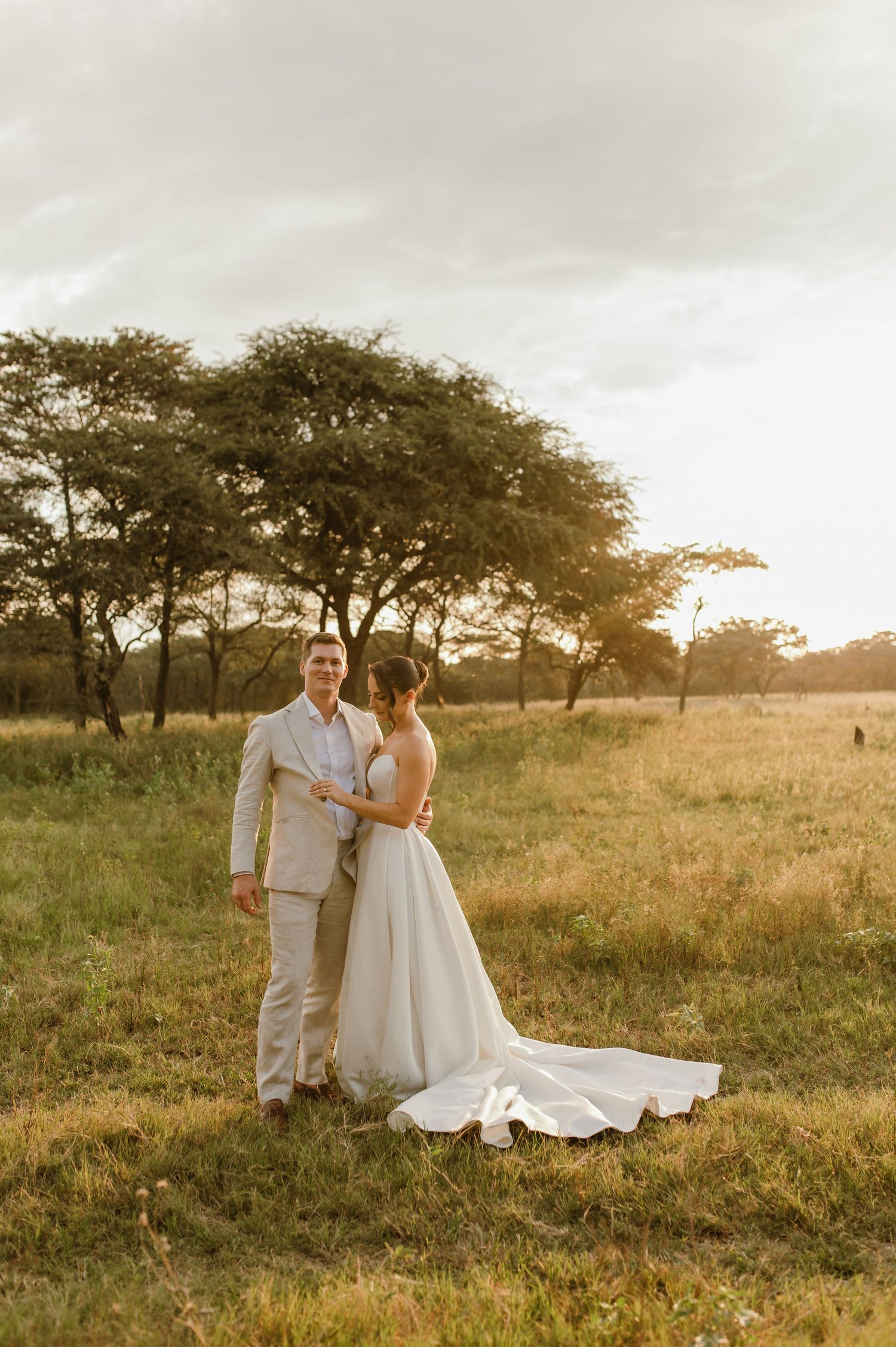 A couple in wedding attire embraces in a golden-lit savanna landscape with acacia trees at sunset.