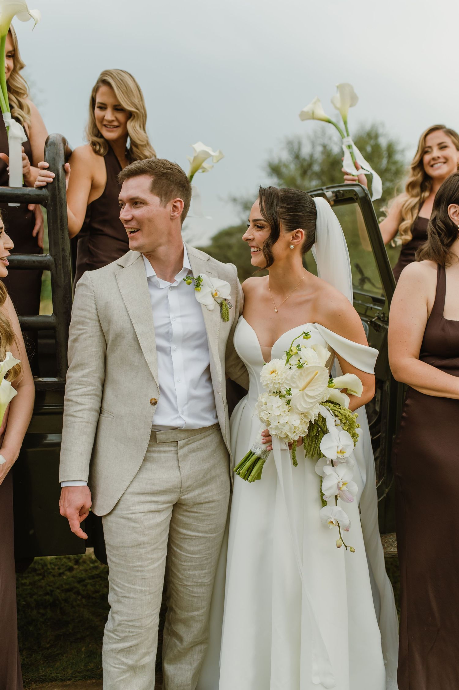 Wedding party in brown dresses and light suits celebrates outdoors with white floral bouquets.
