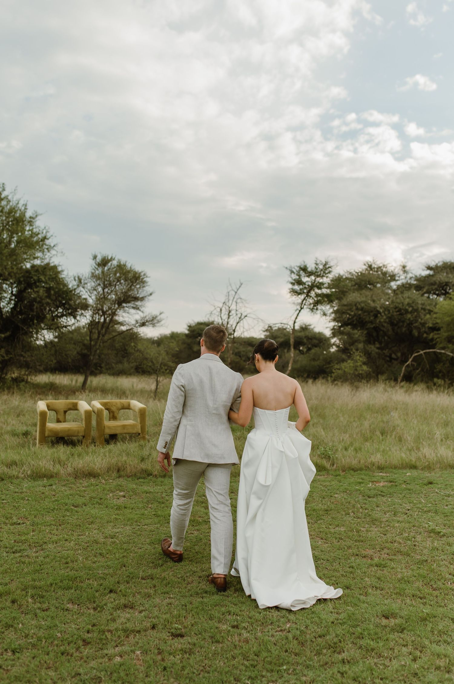 A couple in wedding attire walks hand in hand through a serene grassy field with trees in the background.