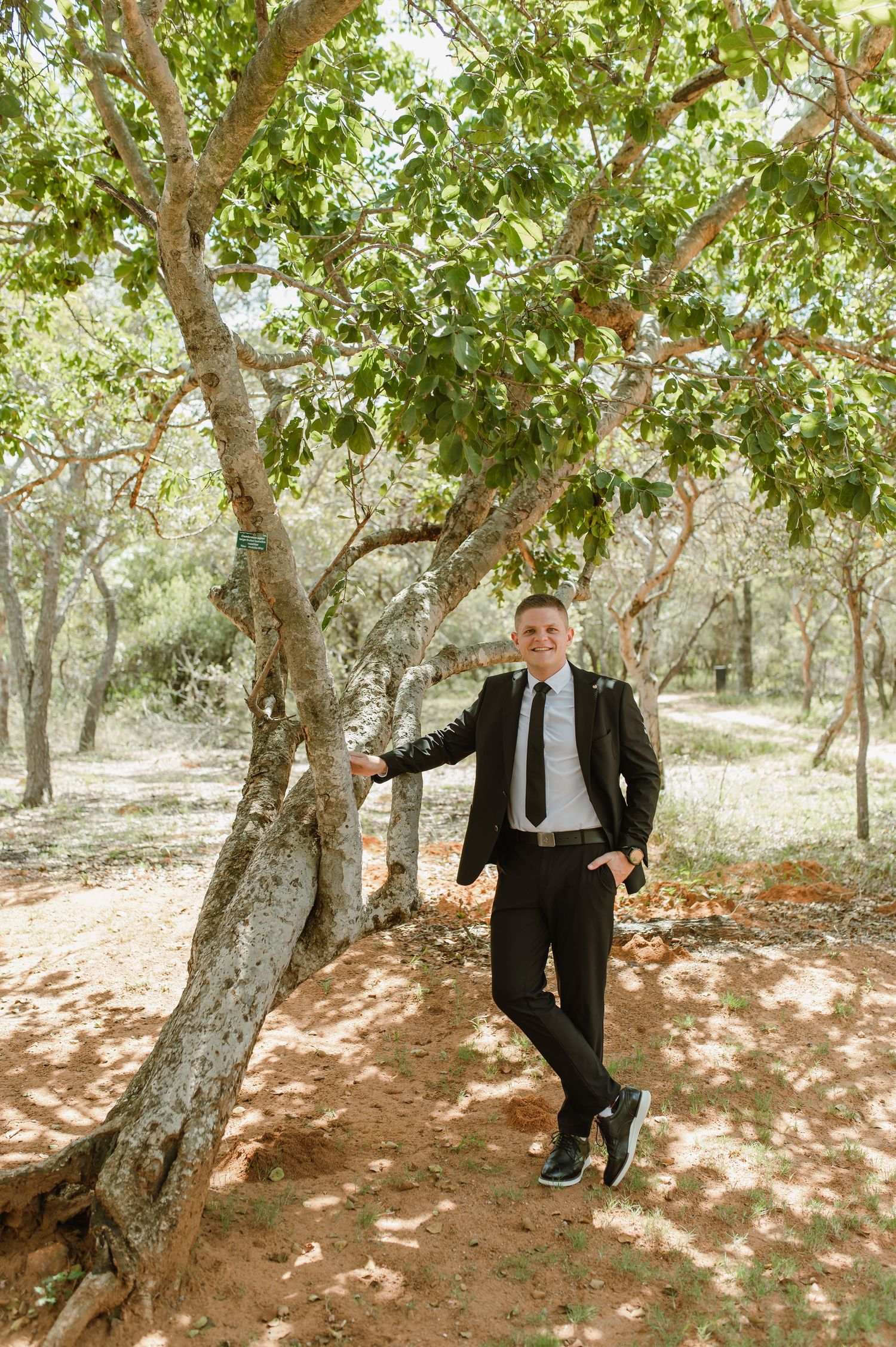 Groom in black suit leaning against a tree in a sunlit woodland setting.