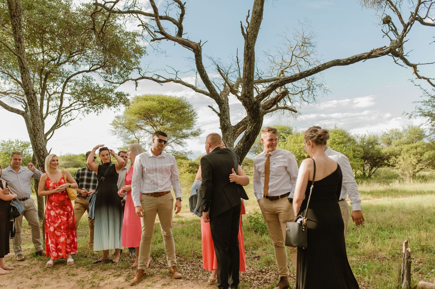 Wedding guests mingle and embrace outdoors surrounded by African bush trees after ceremony celebration.
