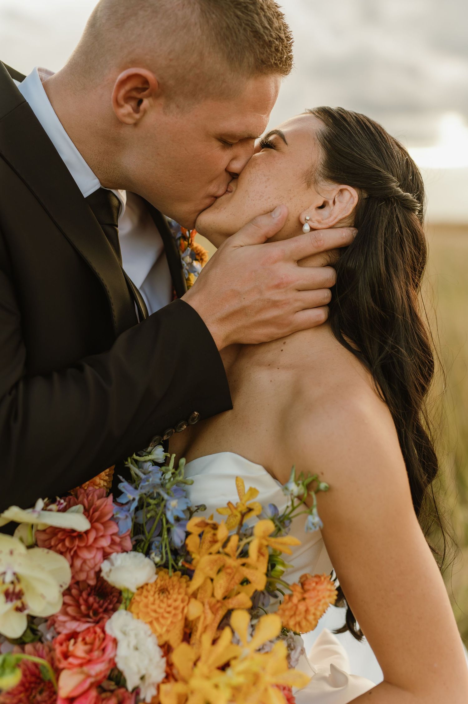 Newlyweds sharing a first kiss with the bride holding a colorful bouquet of dahlias and wildflowers.