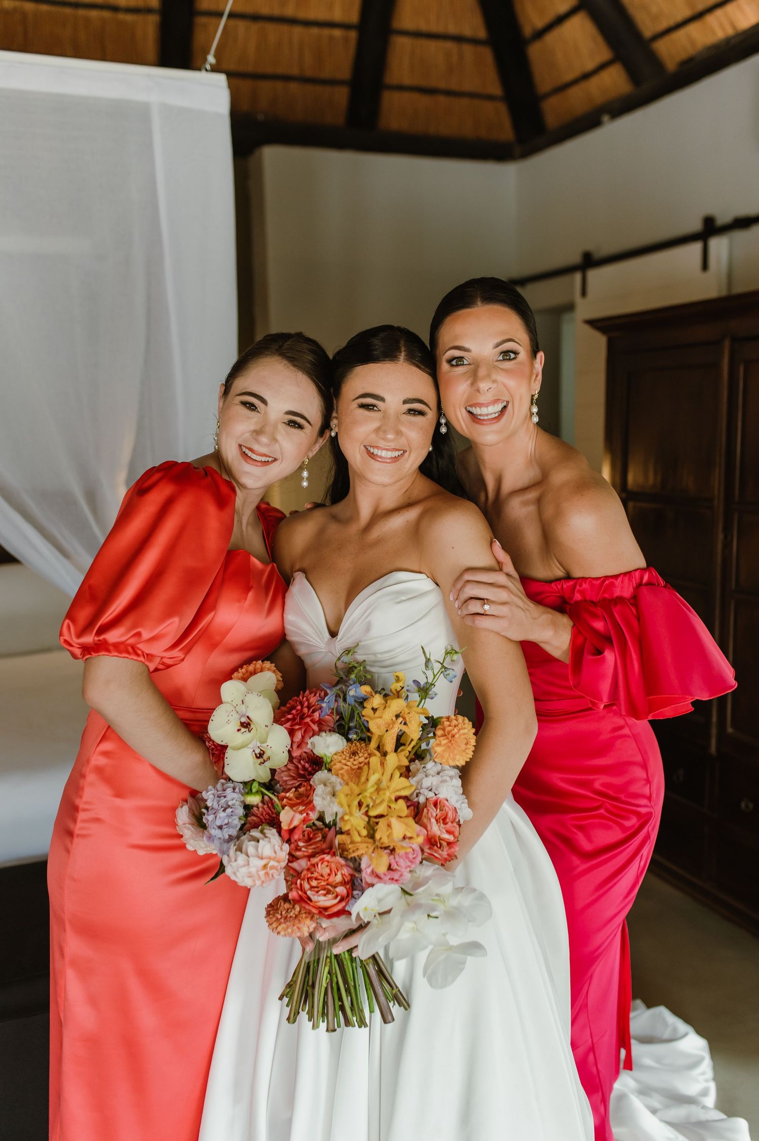 Bride in white gown poses with two bridesmaids in coral and pink dresses holding colorful bouquet.