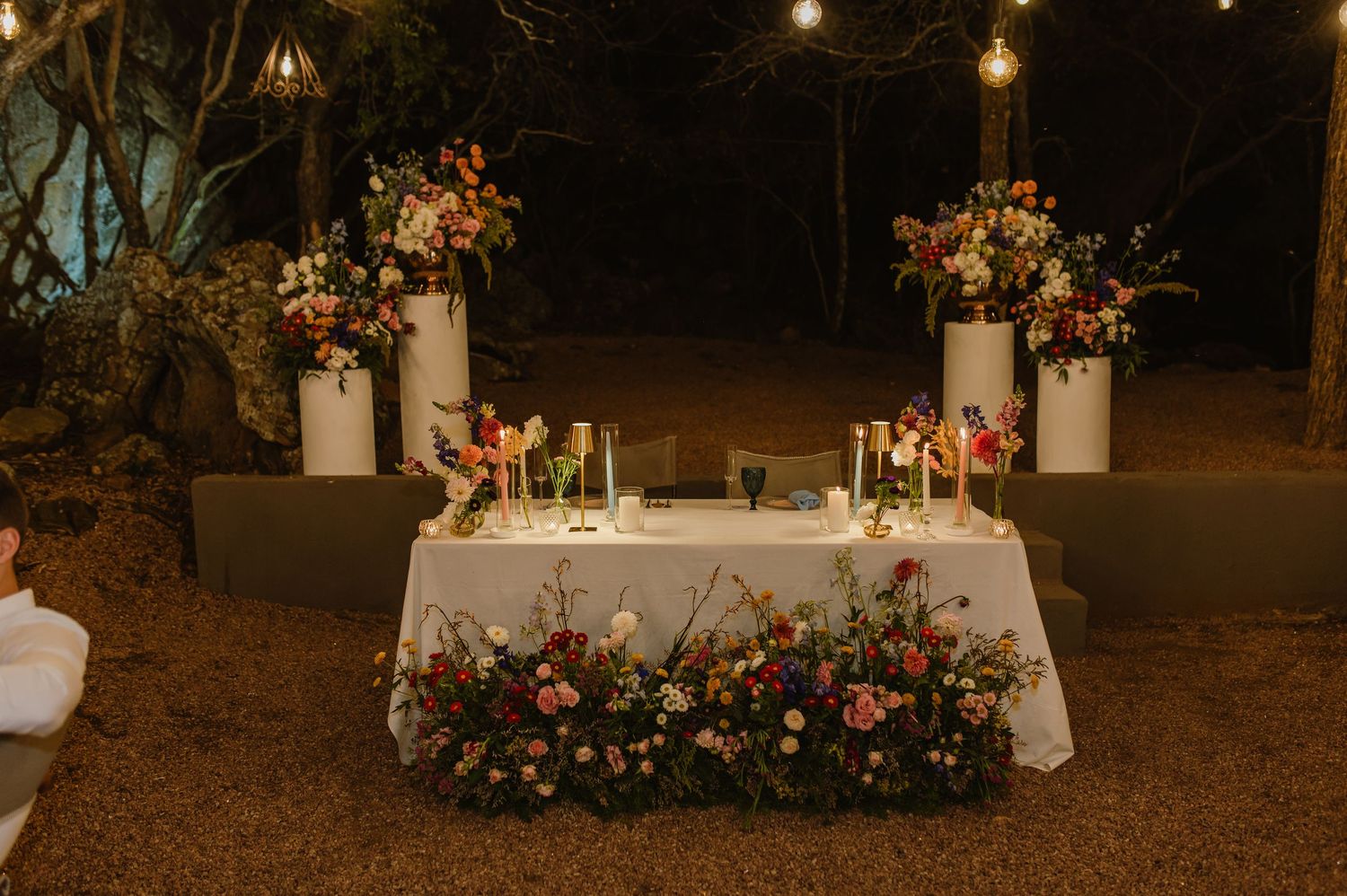 Romantic outdoor sweetheart table adorned with colorful floral arrangements, candles, and string lights at night.