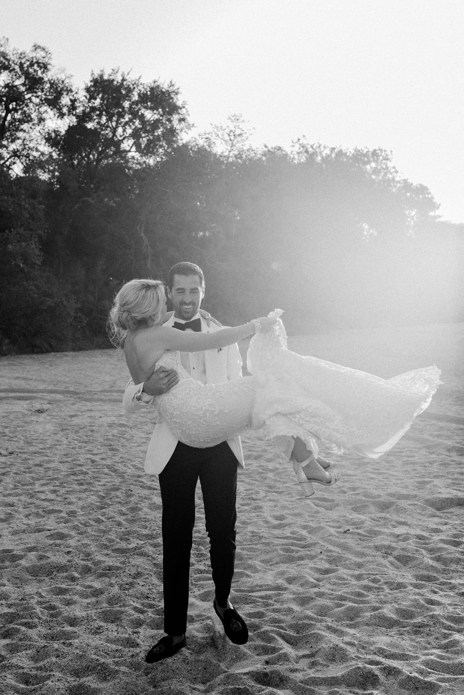 A couple in formal attire dances and spins playfully on a sandy beach at sunset creating a romantic silhouette.