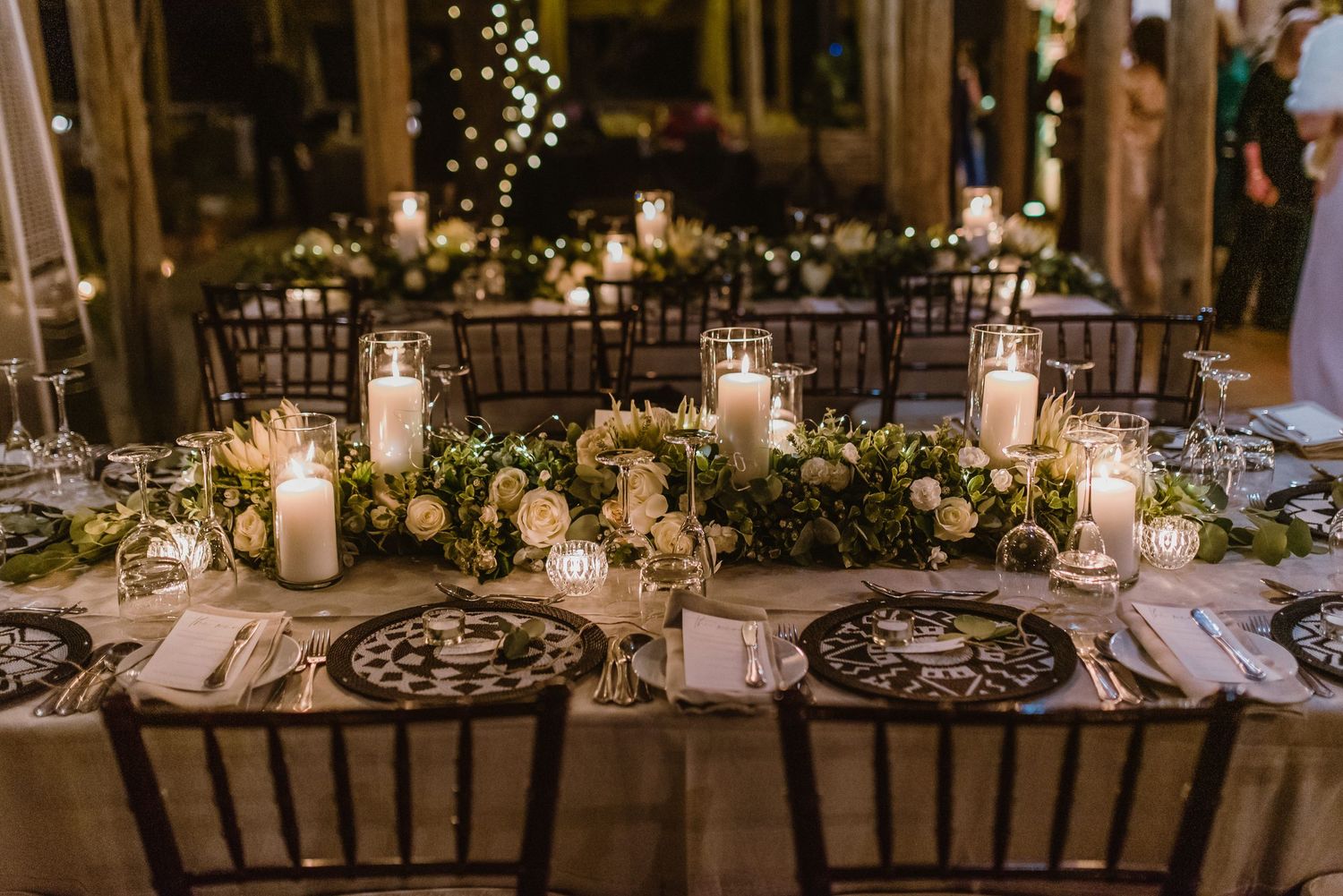 Rustic wedding reception tables decorated with white linens and soft candlelight under wooden beams.