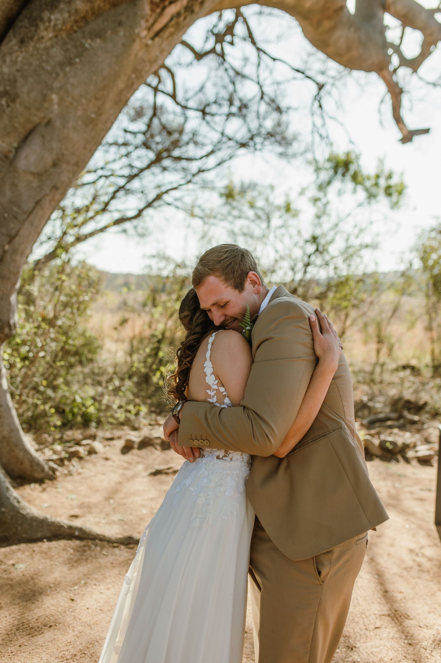A couple embraces during their wedding photoshoot beneath towering trees.
