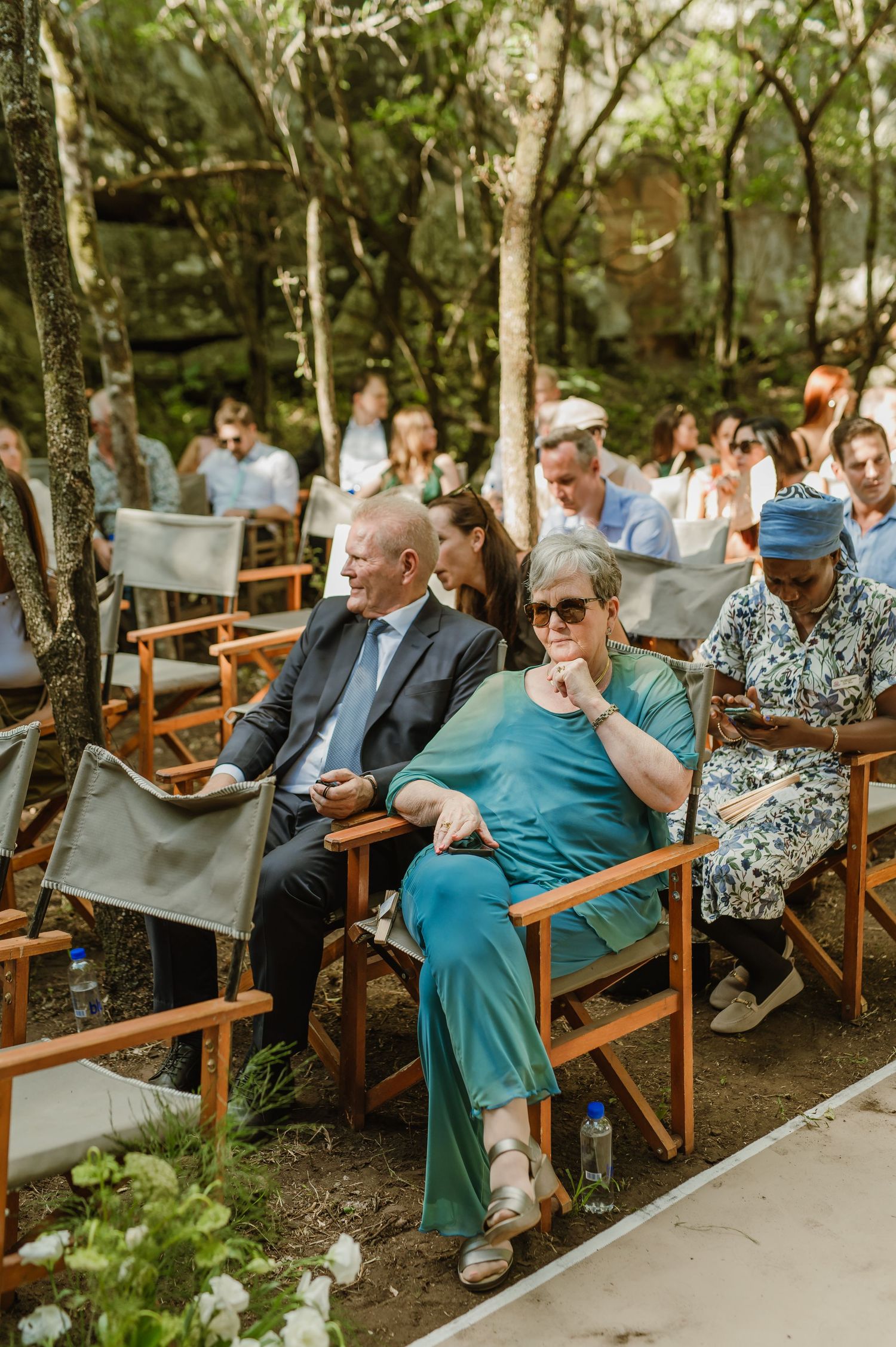Guests in formal attire seated in wooden chairs at an outdoor forest wedding venue.