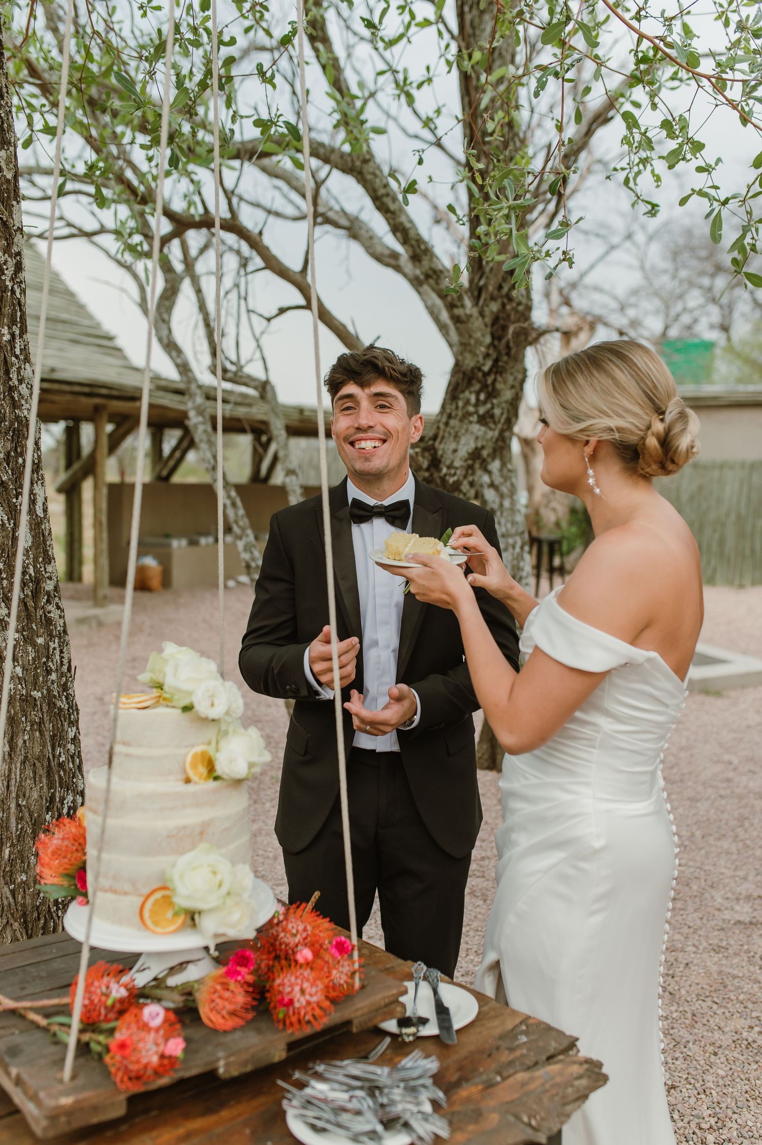 Wedding couple sharing a playful moment while eating cake at an outdoor rustic venue with floral decorations.
