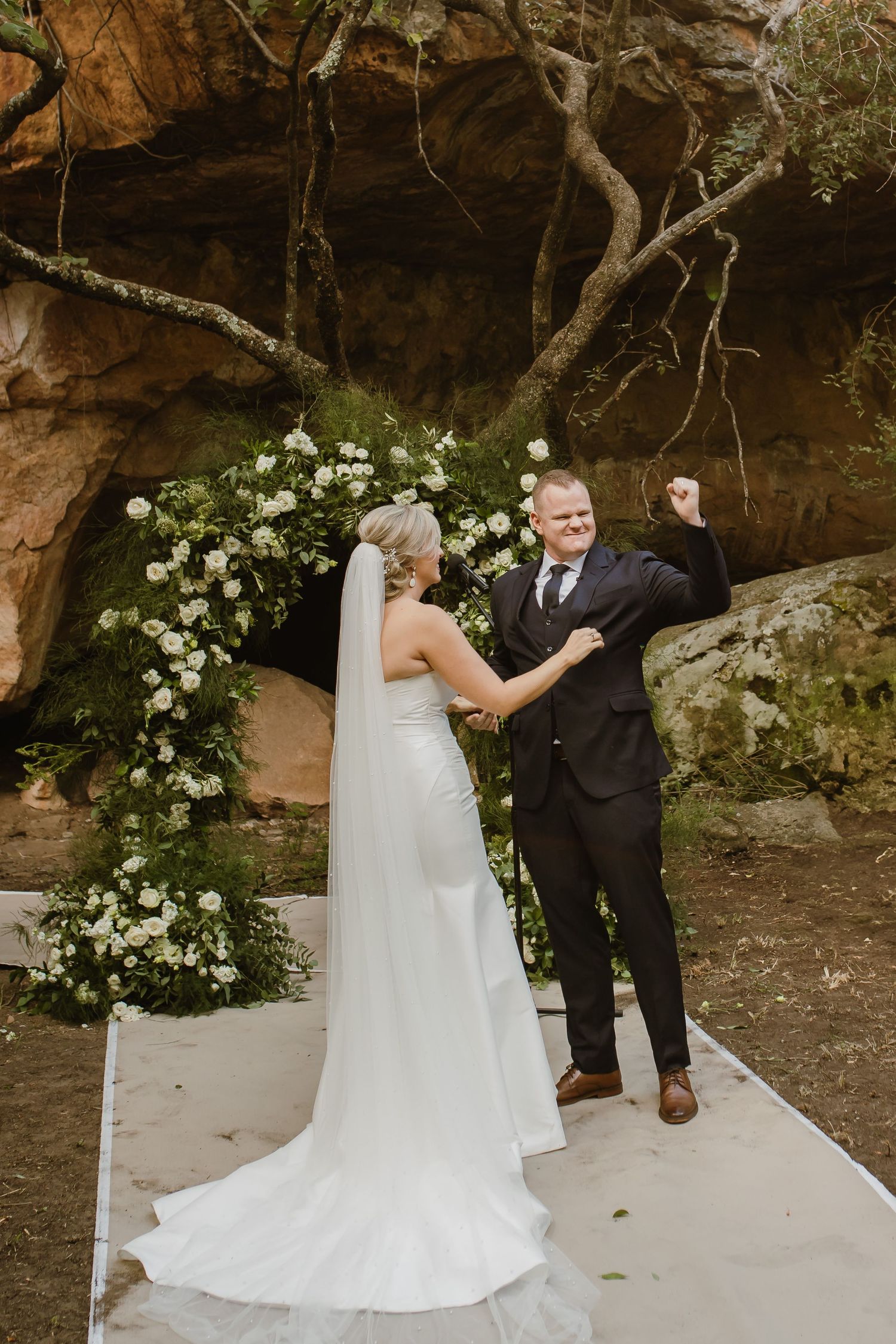 Newlyweds share a sweet first look moment under a white floral arch in a natural cave setting.