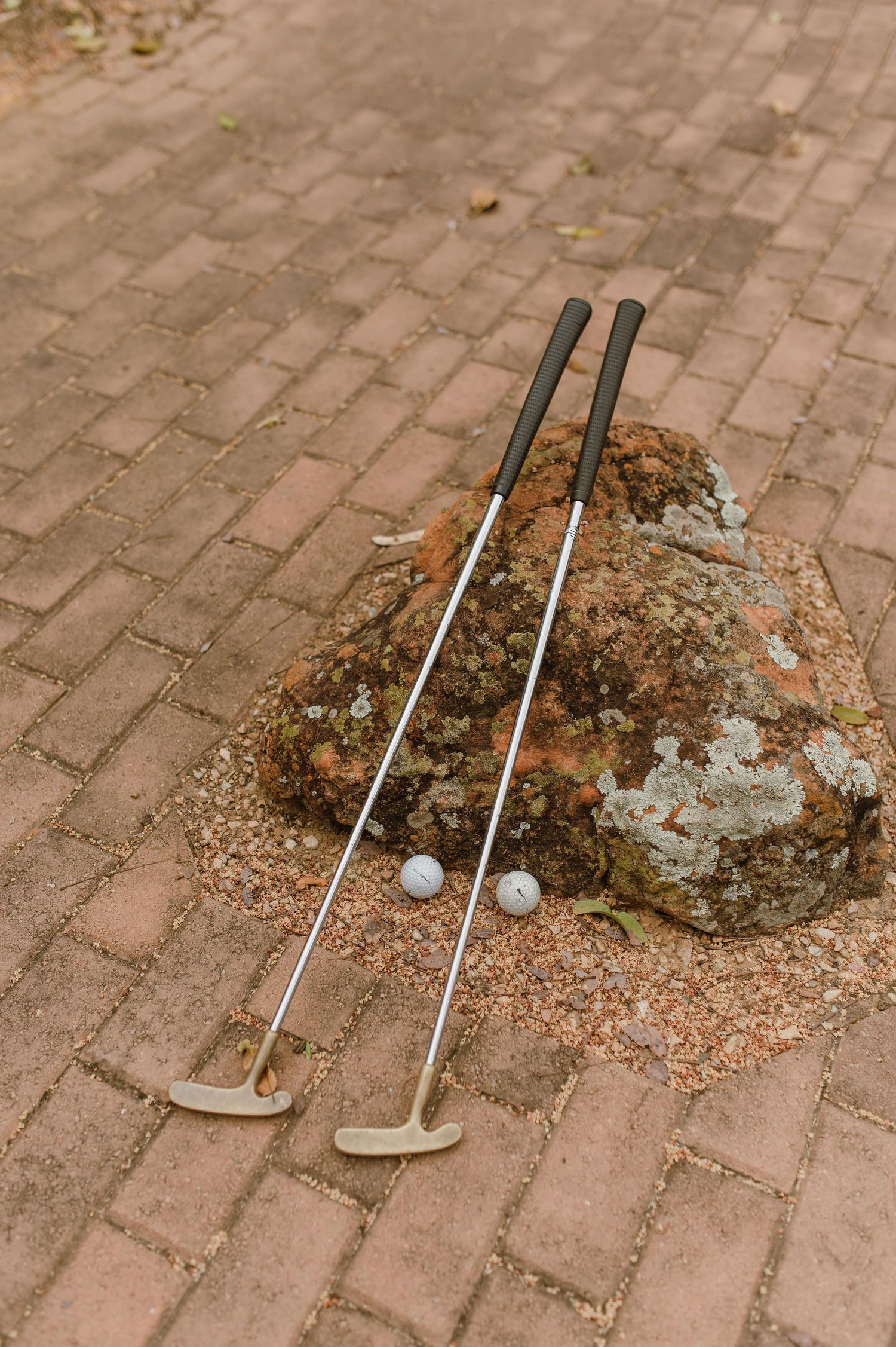 Golf clubs and balls arranged on brick pavers next to a rough patch.