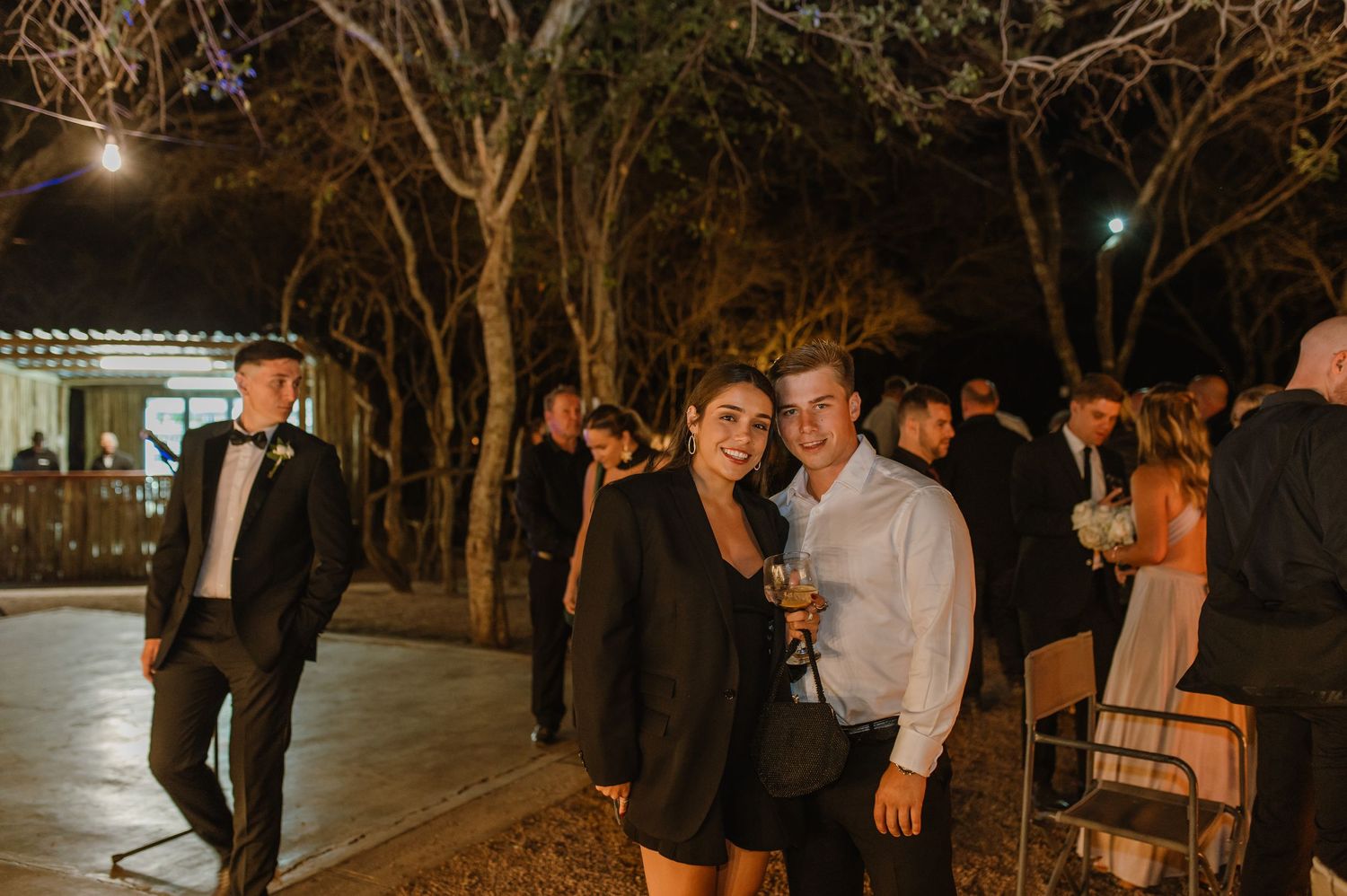 Guests gather beneath illuminated trees at a nighttime outdoor wedding celebration.