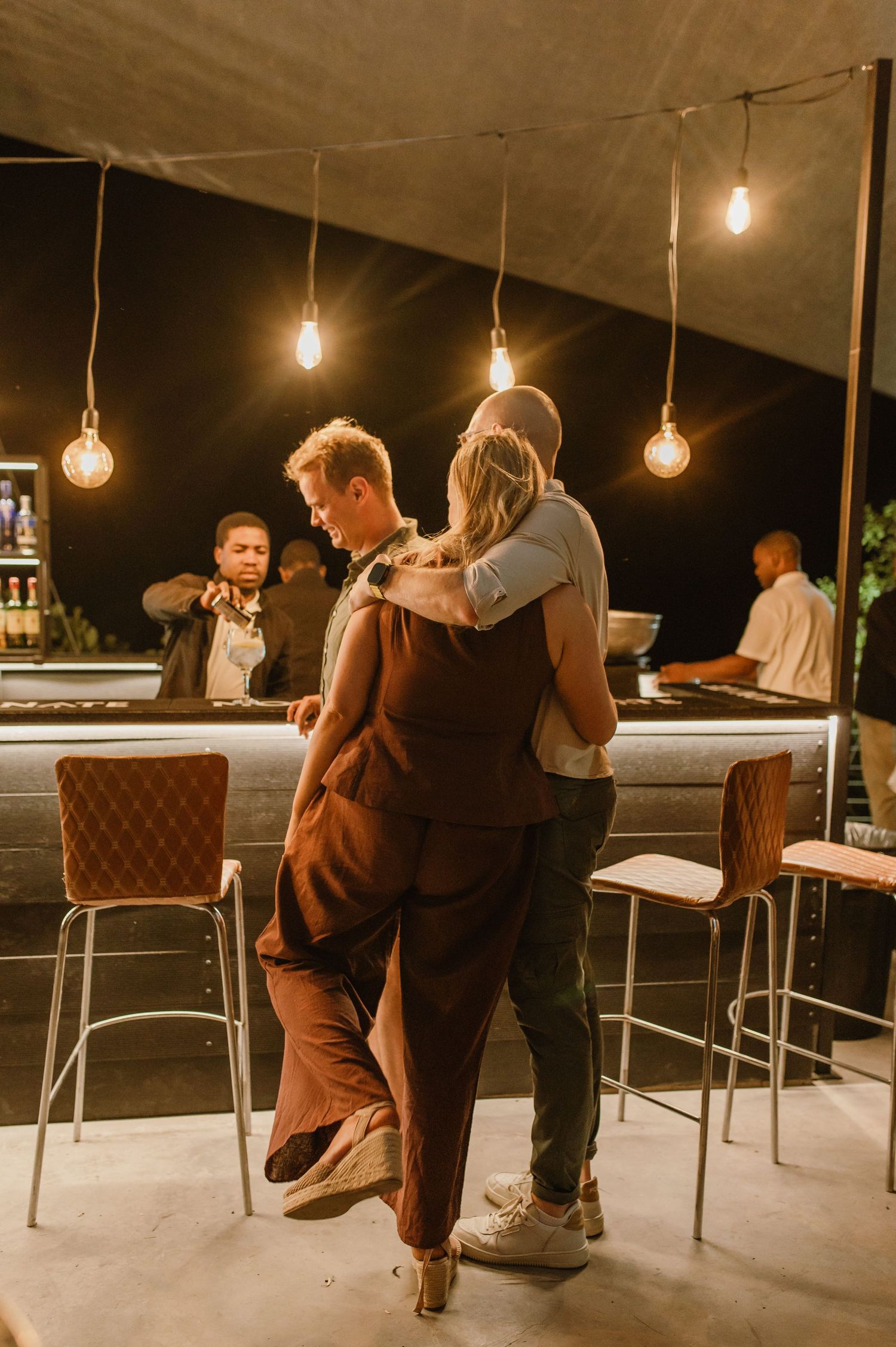 A couple embracing under vintage string lights at a bar with warm ambient lighting.