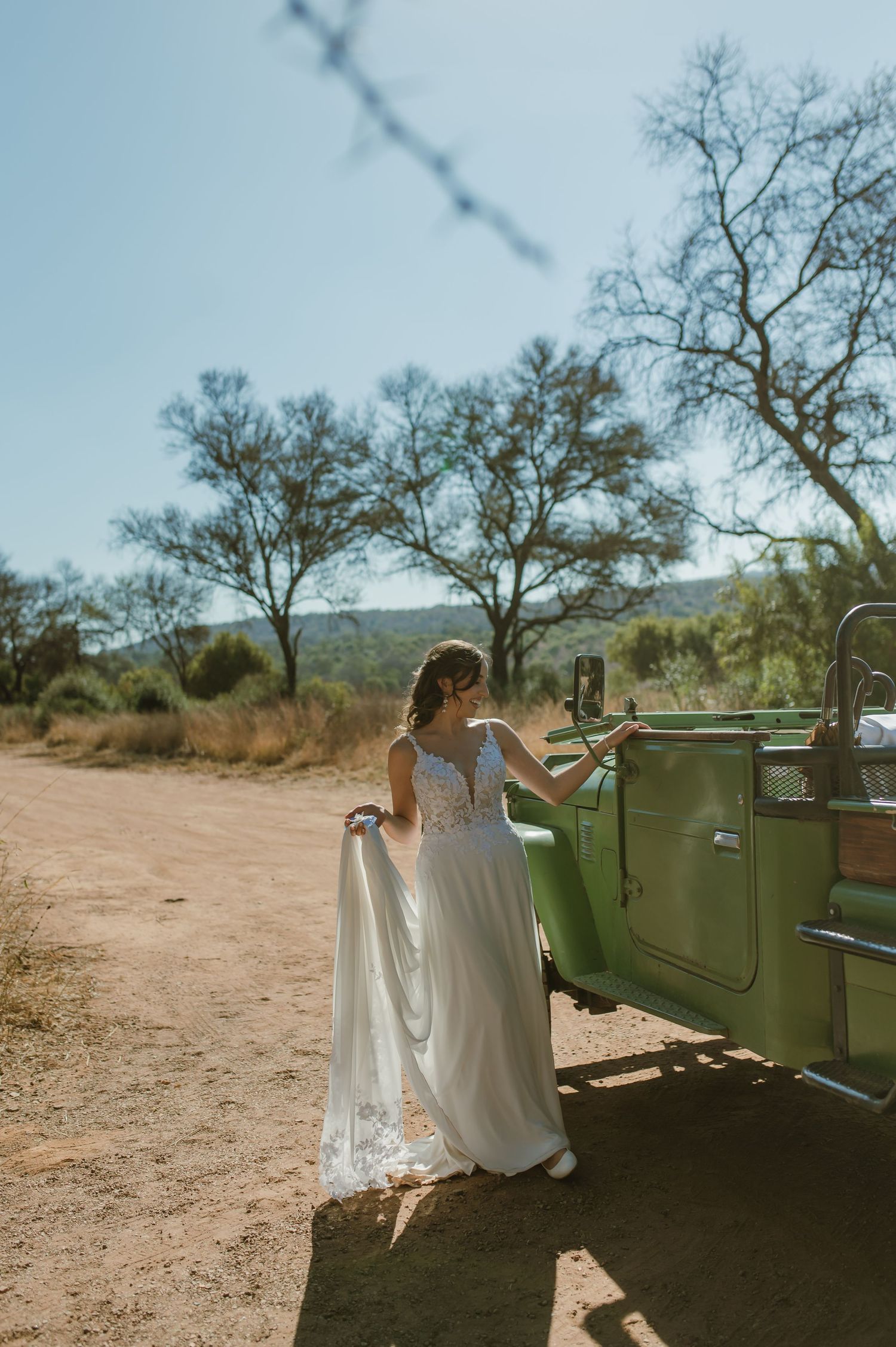 Bride poses playfully next to vintage green vehicle in desert landscape.