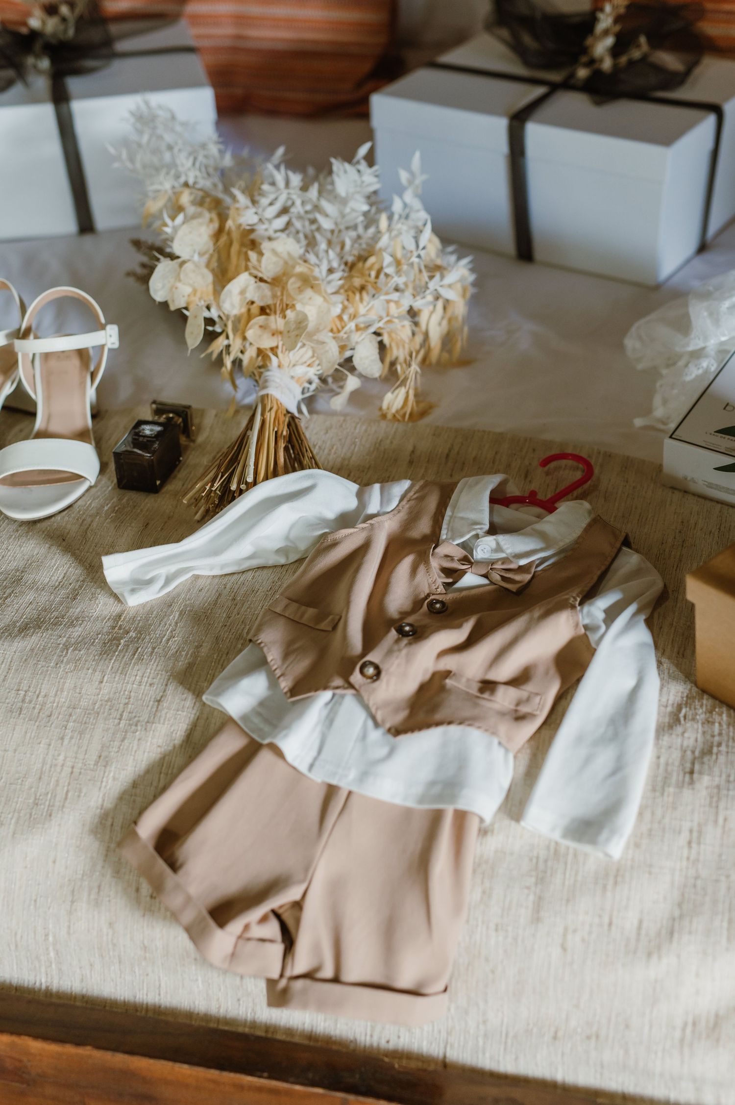 Wedding accessories and clothing laid out on neutral linen including dried flowers and fabric.