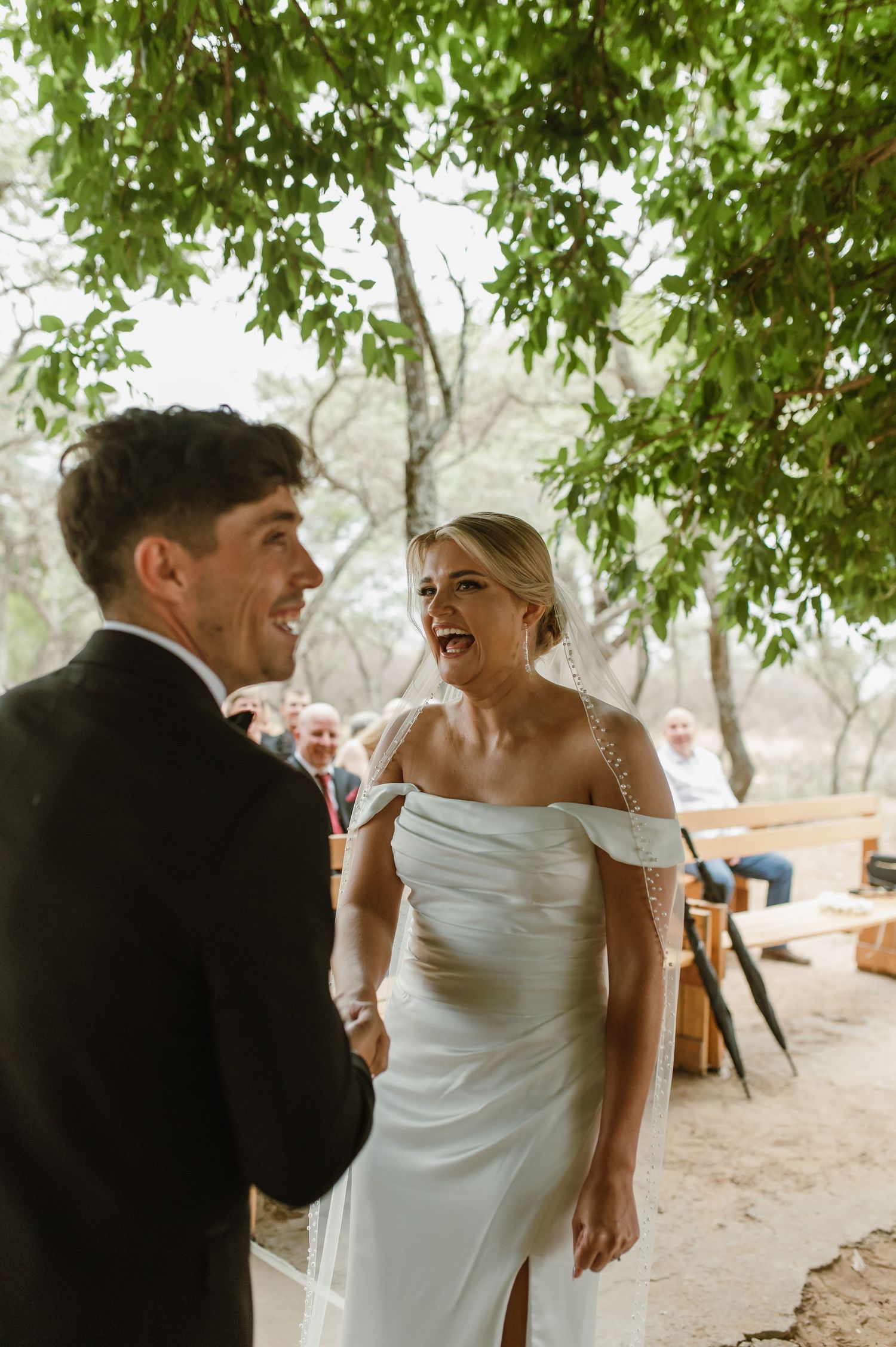 A joyful wedding moment captured under green trees with guests seated on wooden benches in the background.