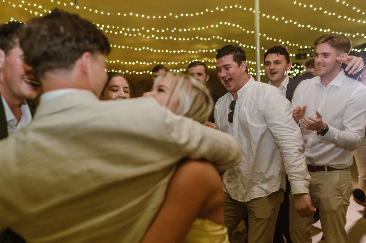 Wedding guests dance and celebrate under warm string lights at an evening reception.