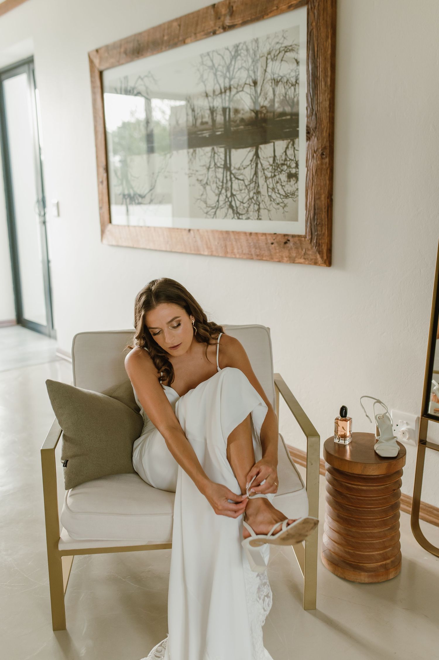 A bride in a white gown sits on a chair putting on shoes with a framed artwork behind her.