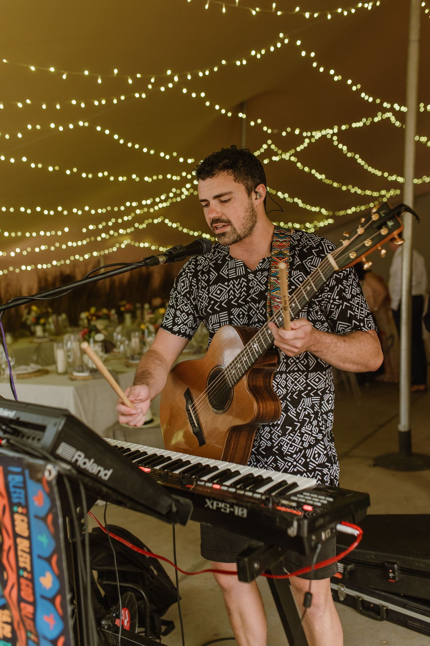 Musician performs with guitar and keyboard under string lights at an outdoor wedding reception.
