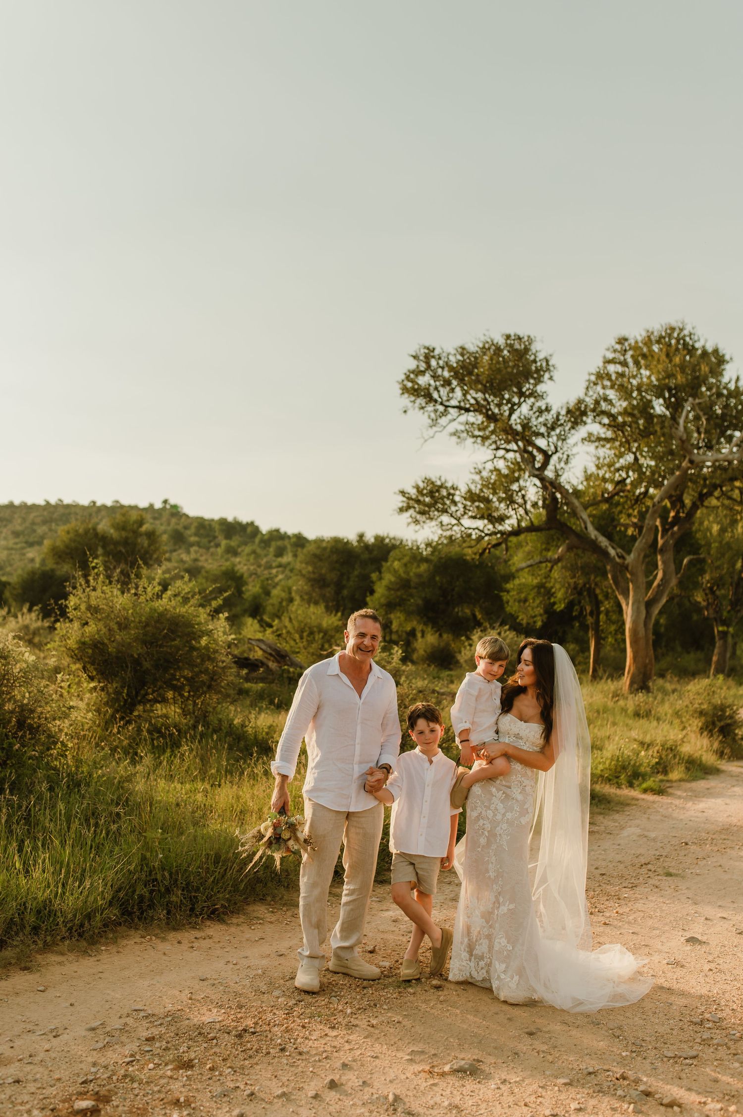 A joyful wedding family portrait on a sandy path surrounded by lush greenery at golden hour.
