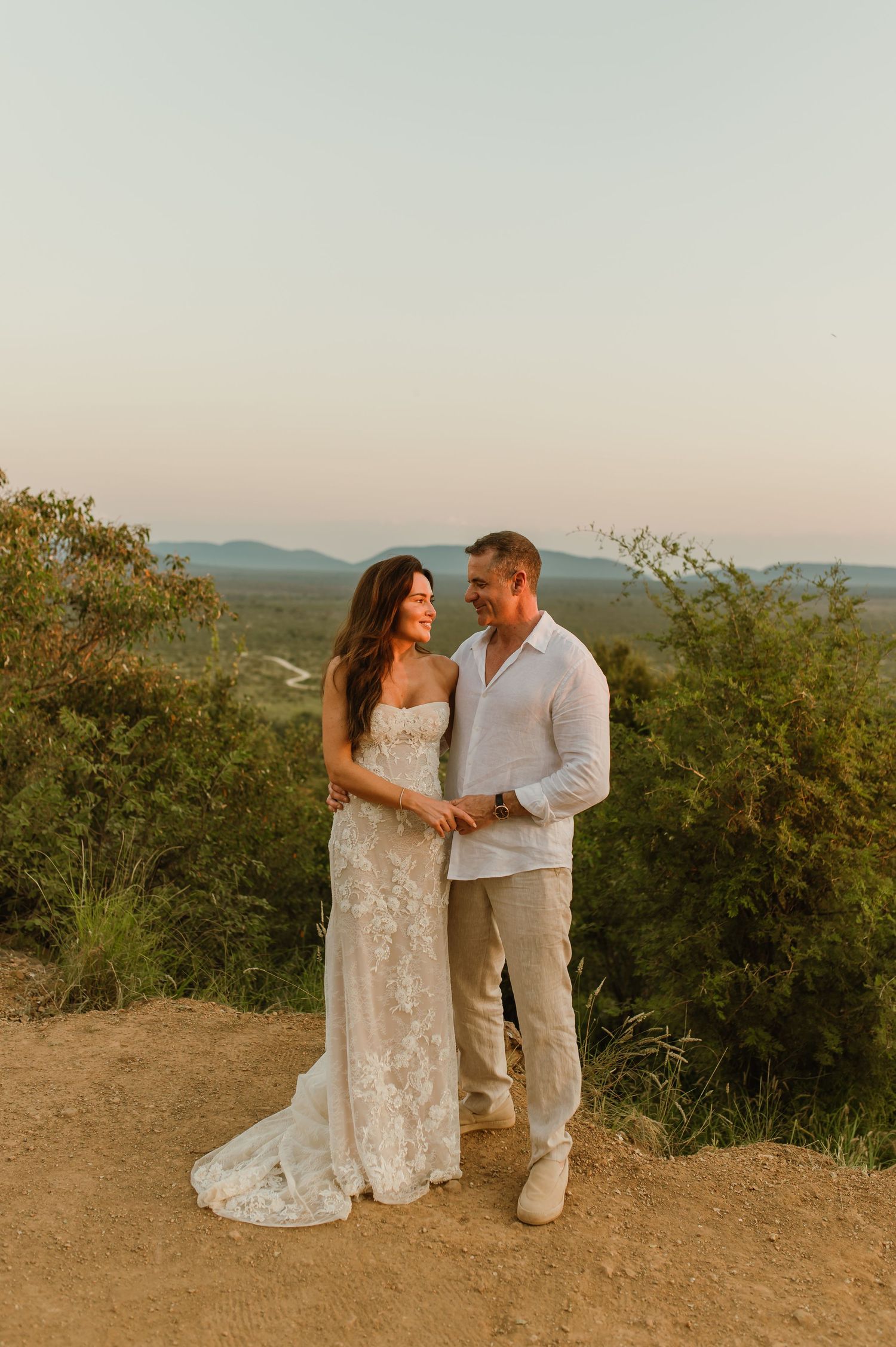 Bride in lace gown and groom in linen smiling together at sunset with scenic valley view.