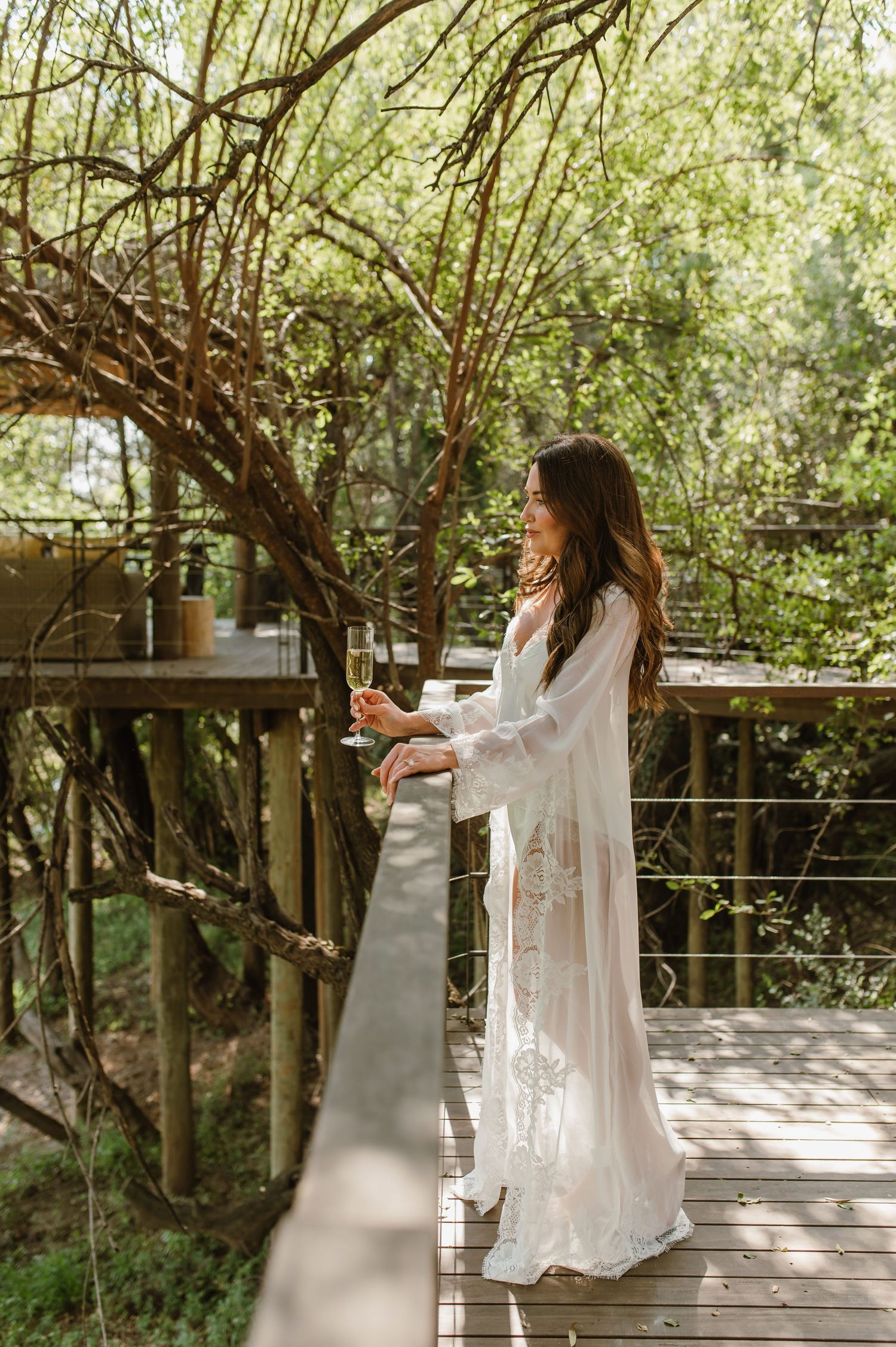 Bride in white lace robe holding champagne on wooden treehouse deck surrounded by lush green trees.