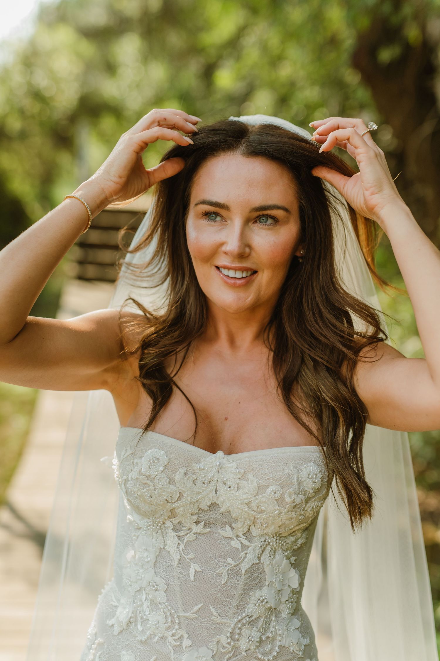 A beautiful bride in a strapless lace wedding dress adjusts her veil outdoors among green trees.